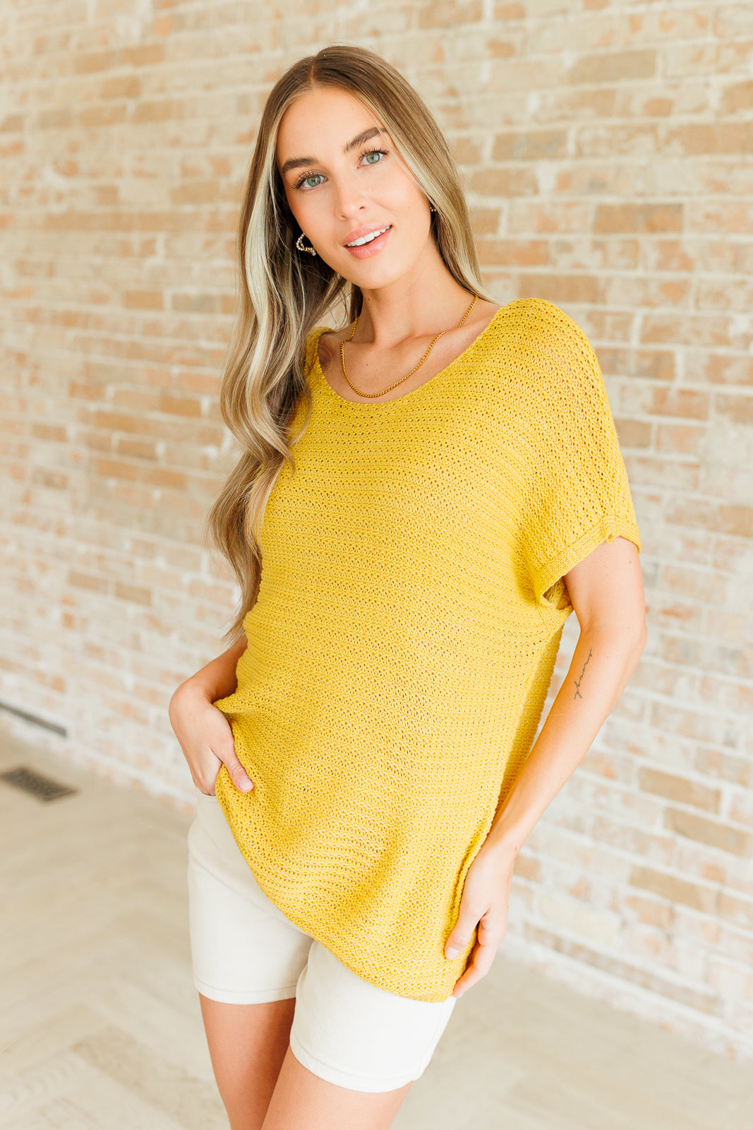 Woman wearing loose yellow knit top with short sleeves and beige shorts standing indoors against light brick wall