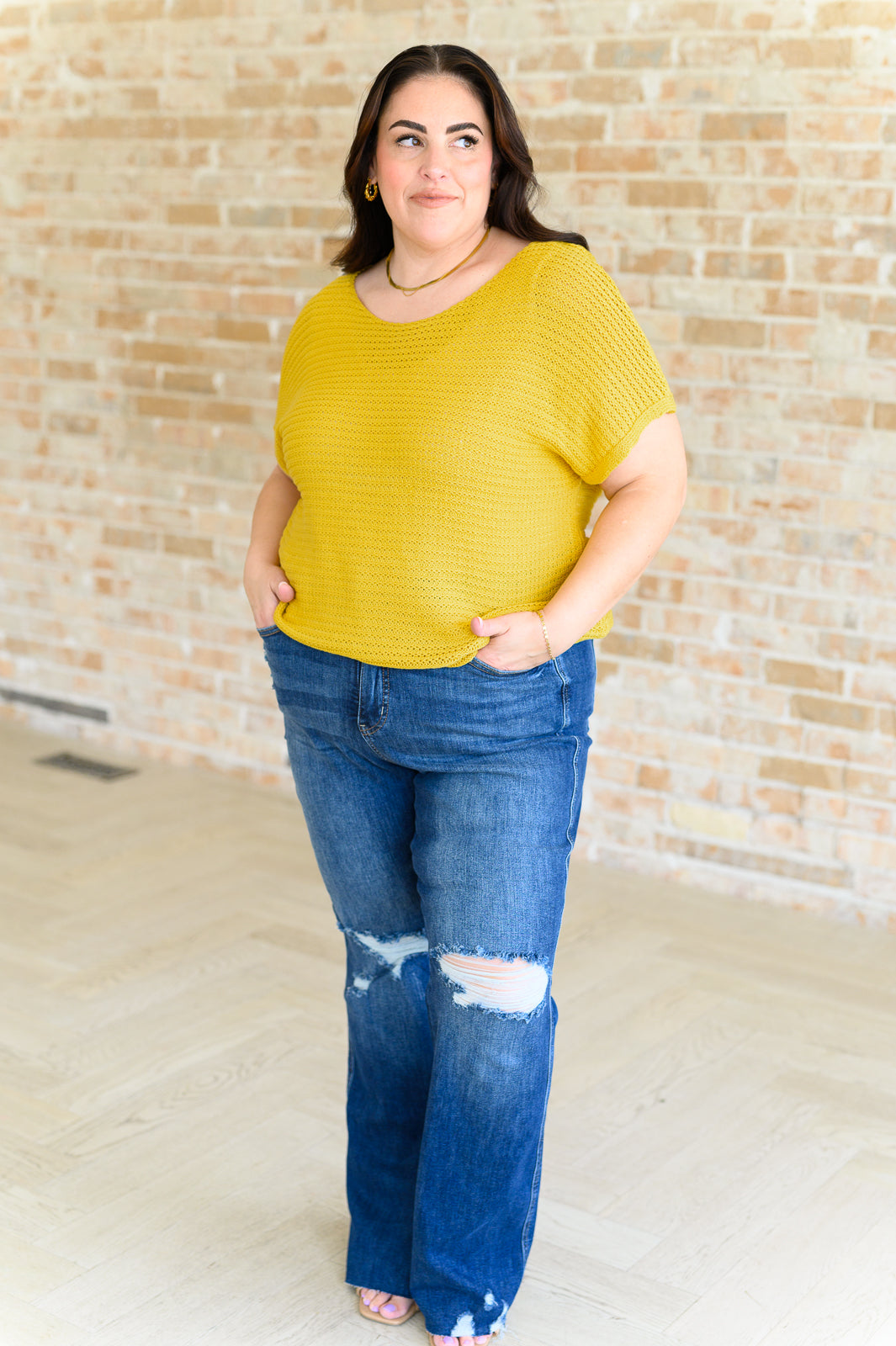 Plus size woman wearing textured yellow knit top with short sleeves and distressed jeans standing indoors against brick wall