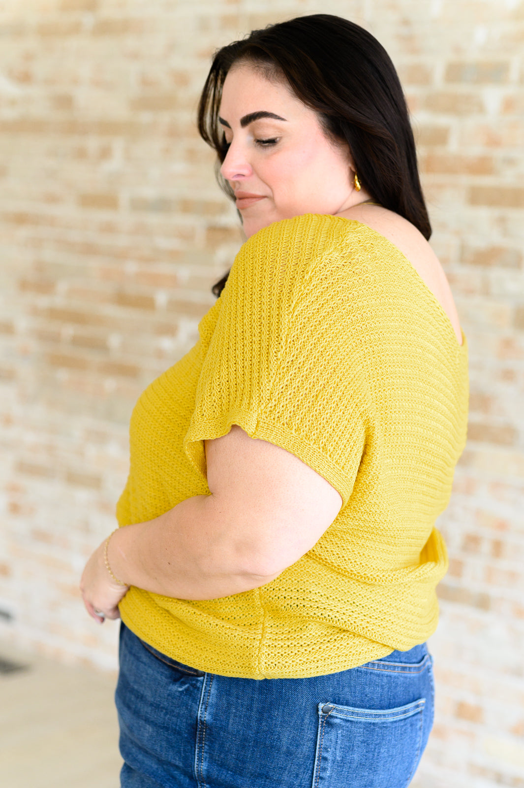 Plus size woman wearing bright yellow knit top with short sleeves and scooped back neckline against beige brick wall