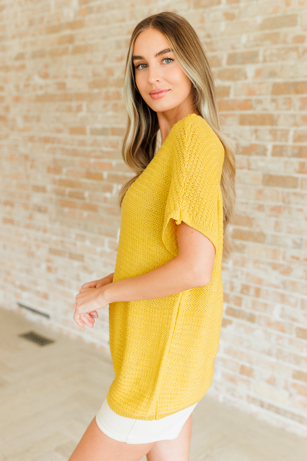 Side view of woman wearing textured yellow knit top with short sleeves and casual fit against brick wall background