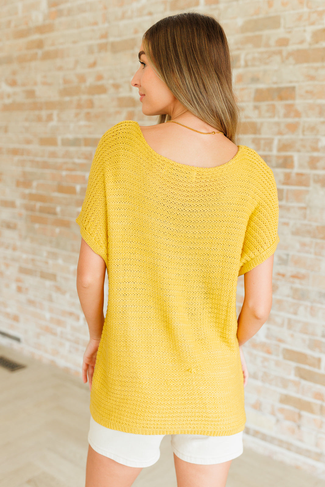 Woman shown from behind wearing bright yellow knit top with short sleeves and scooped neckline against brick wall