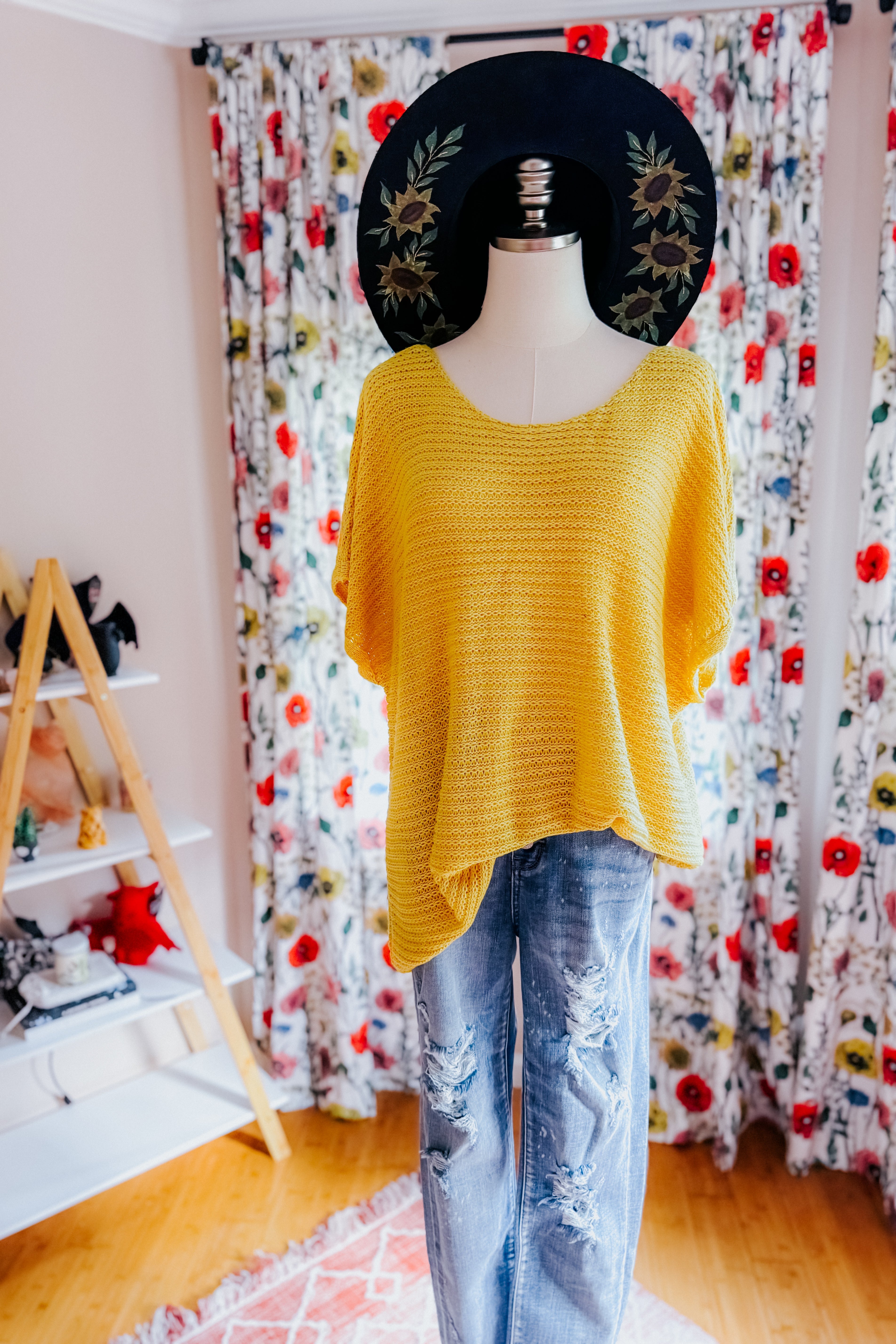Mannequin dressed in loose yellow knit top with wide neckline and distressed blue jeans indoors with floral curtains and hat