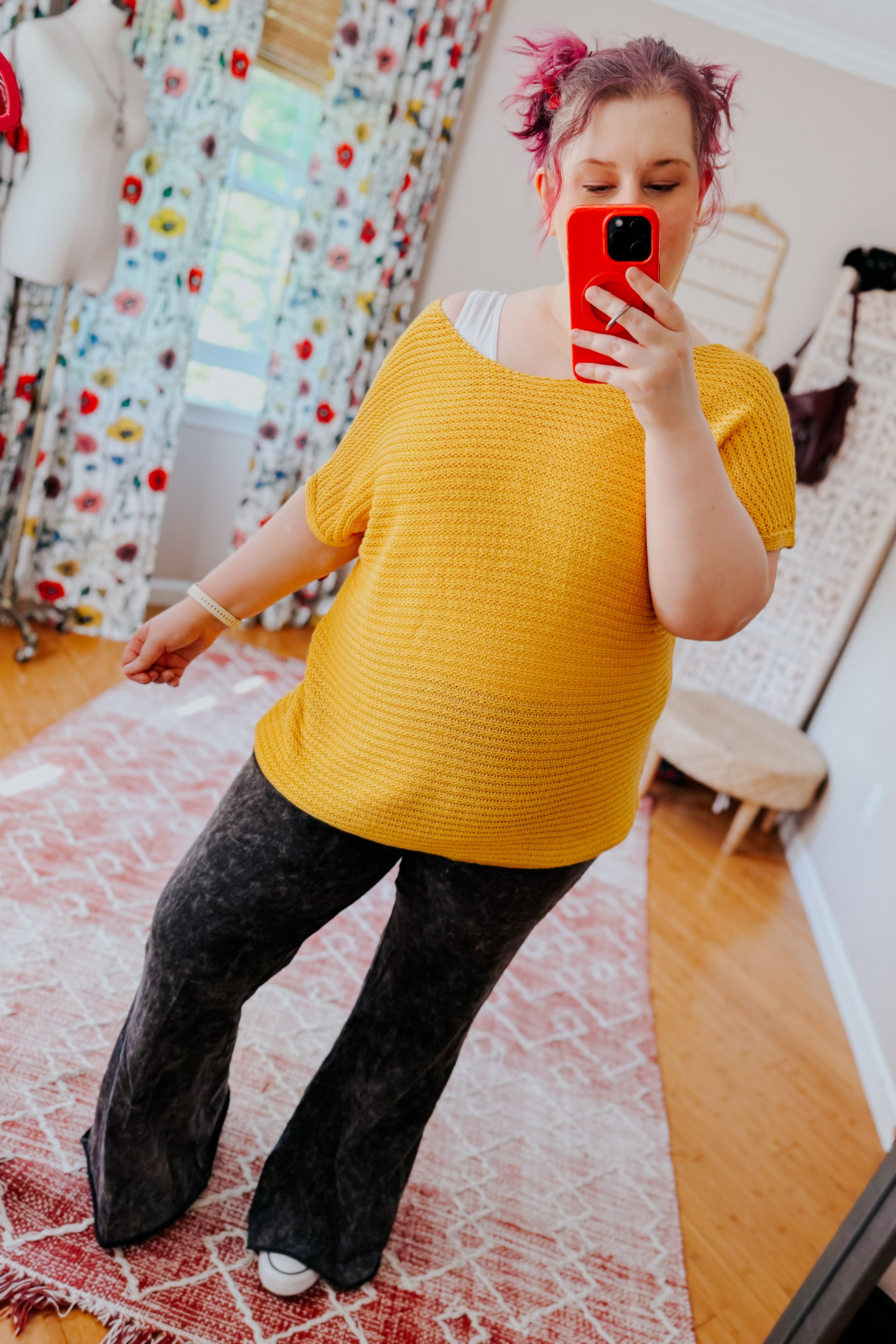 Woman taking mirror selfie wearing loose yellow knit top with short sleeves paired with black flared pants indoors