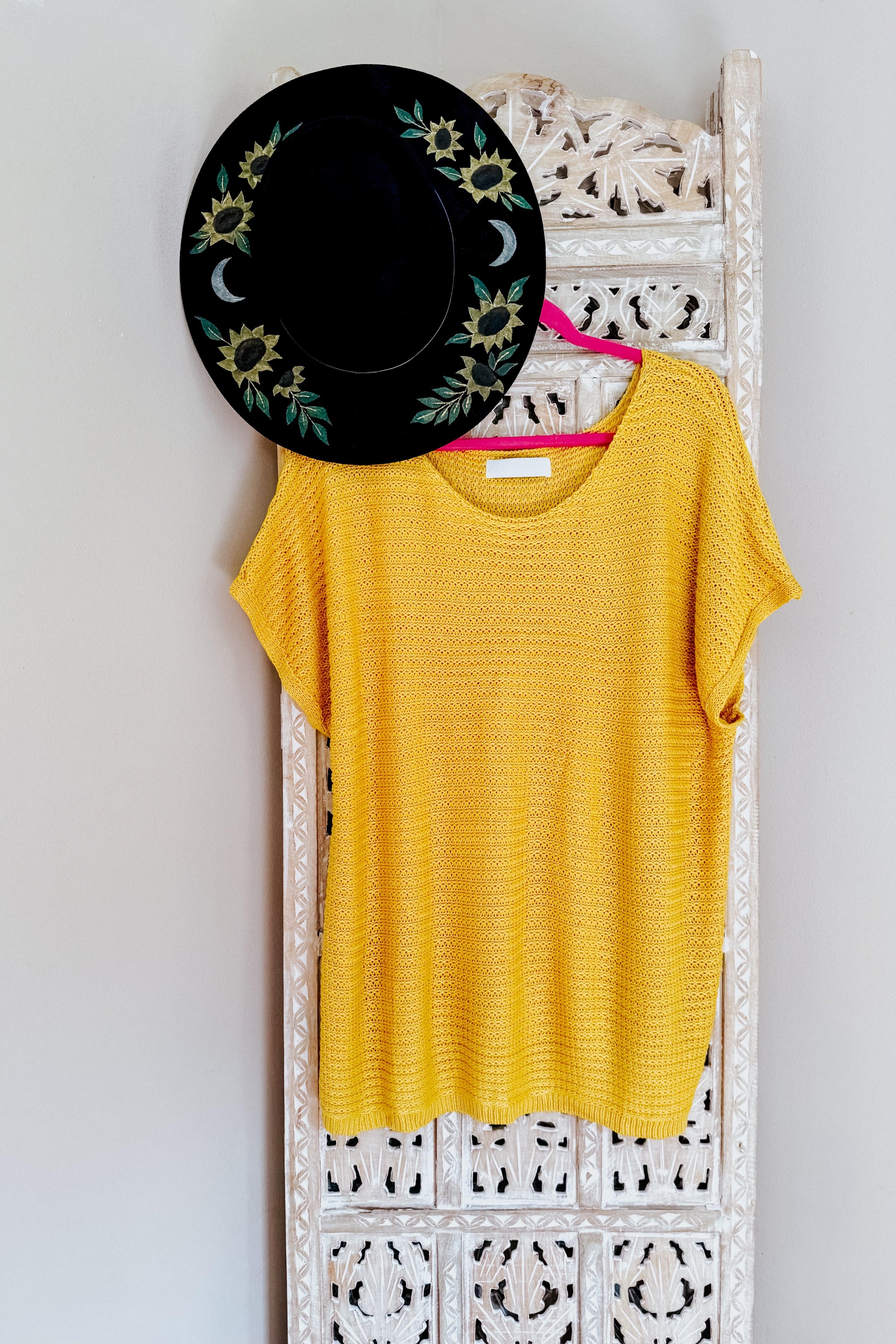 Yellow knit top with short sleeves hanging on carved white wooden screen with black embroidered hat above