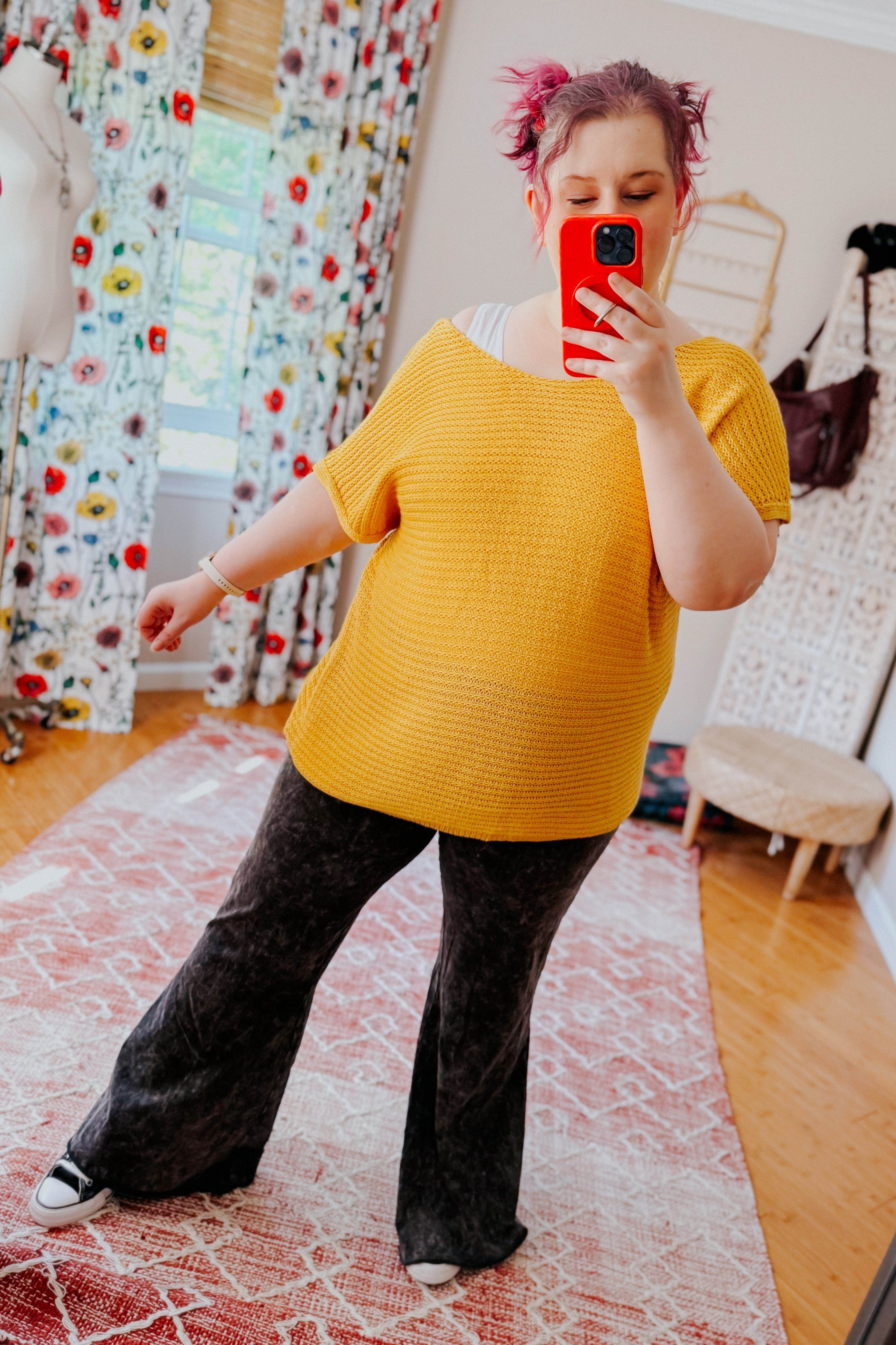 Woman taking mirror selfie wearing loose yellow knit top with short sleeves paired with black pants in colorful room