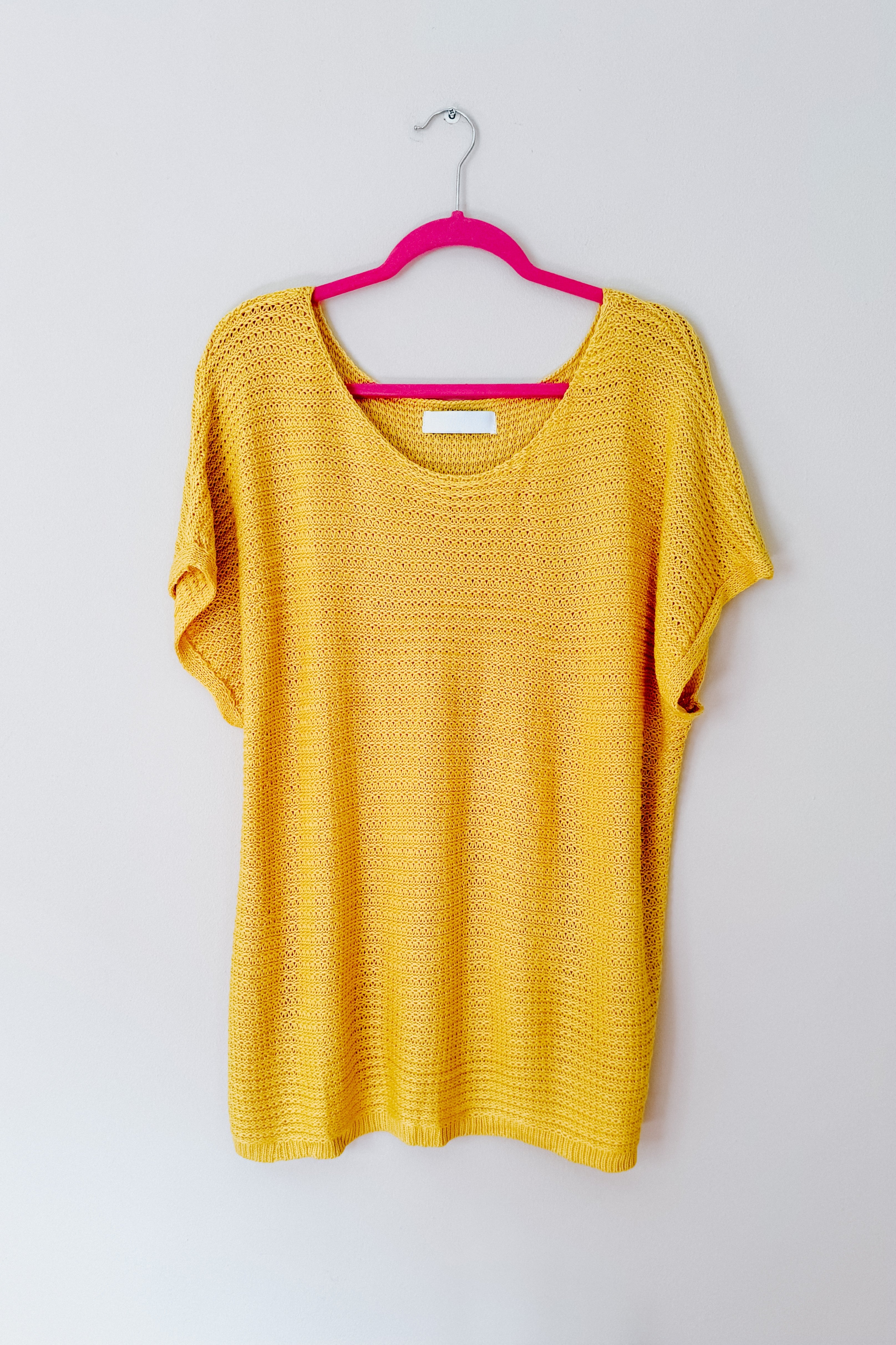 Lightweight yellow knit top with short sleeves and scoop neckline hanging on a pink hanger against plain wall
