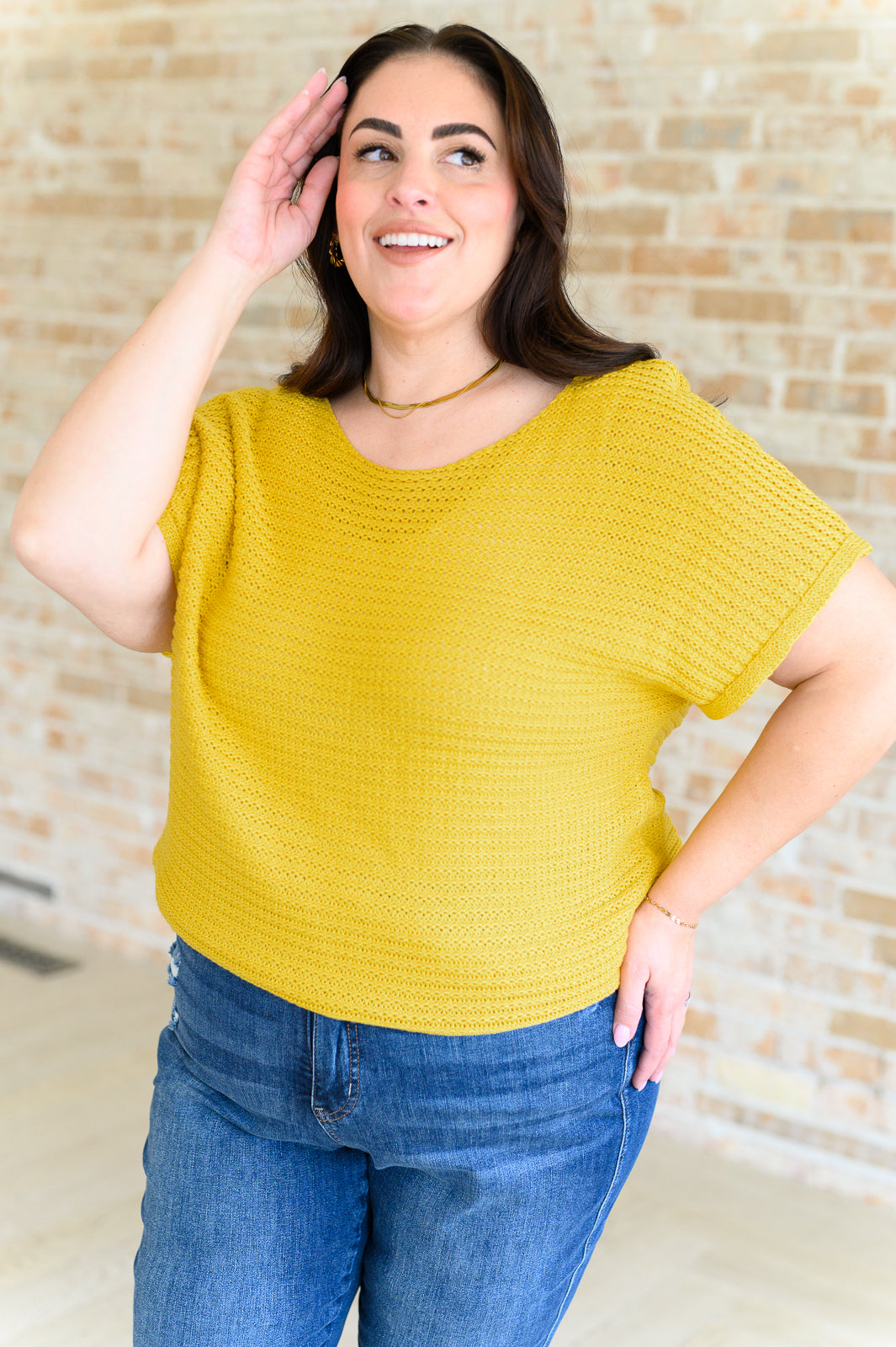 Smiling woman wearing loose yellow knit top with short sleeves and blue jeans standing indoors against brick wall