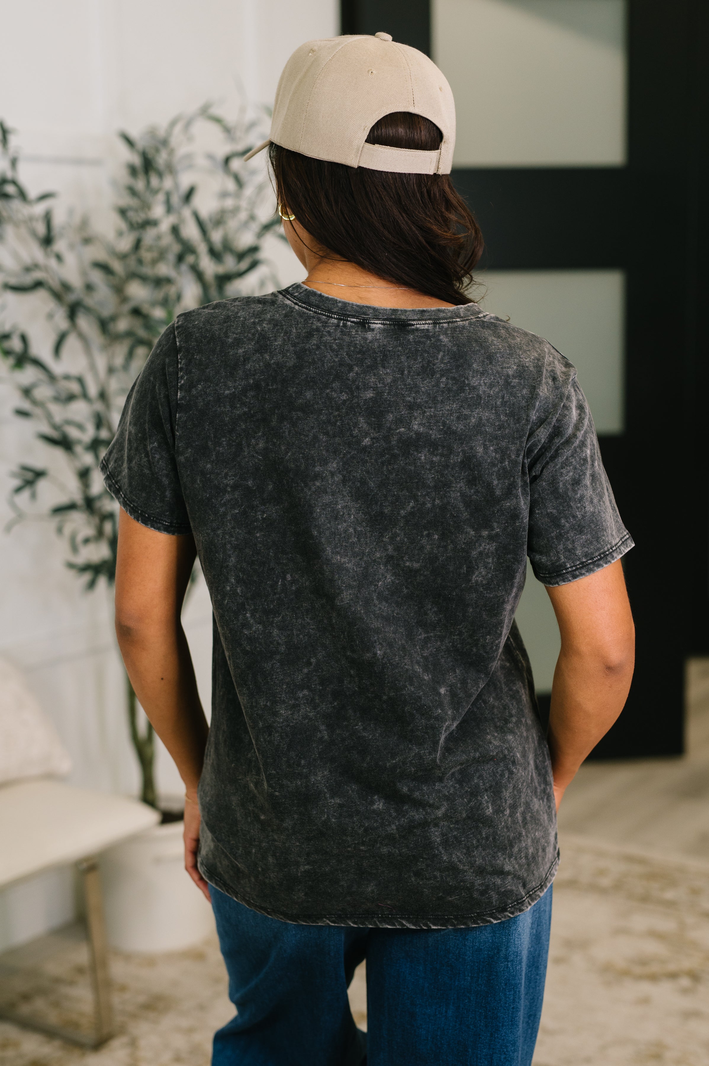 Back view of person wearing ash black mineral wash tee paired with blue jeans and beige cap indoors