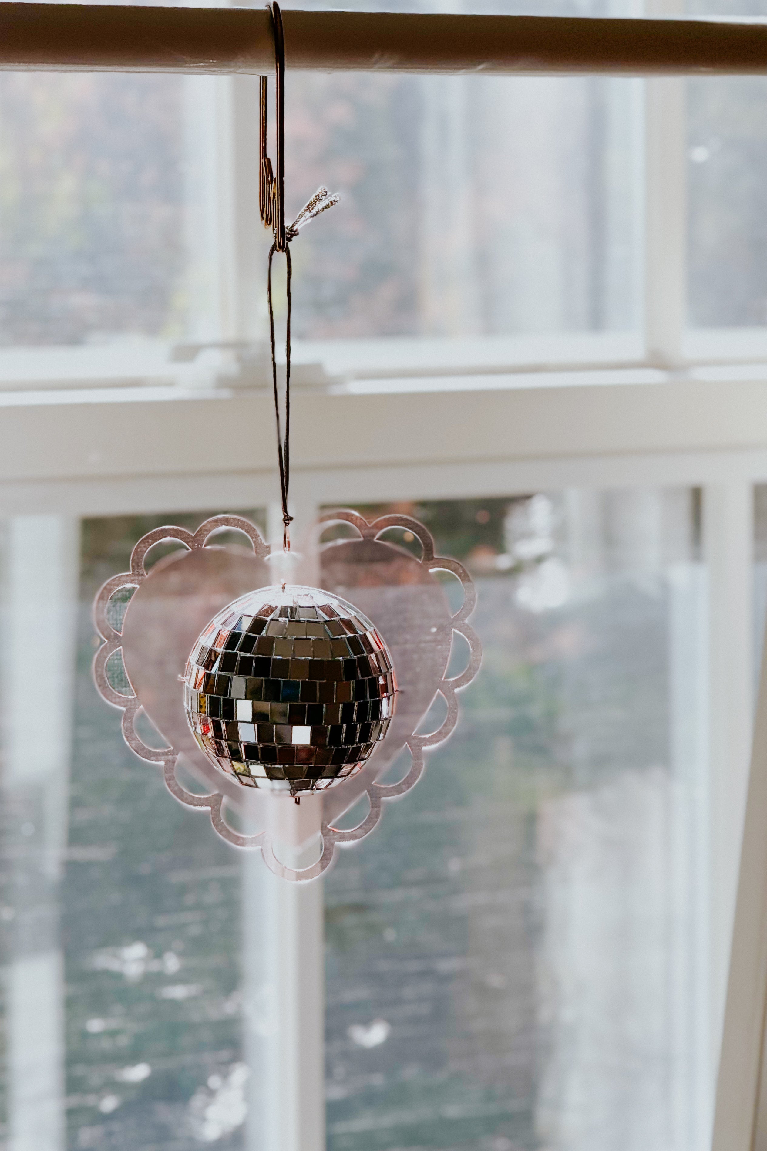 Soft pink scalloped heart sun catcher featuring a reflective mini disco ball hanging by black string before windowpane