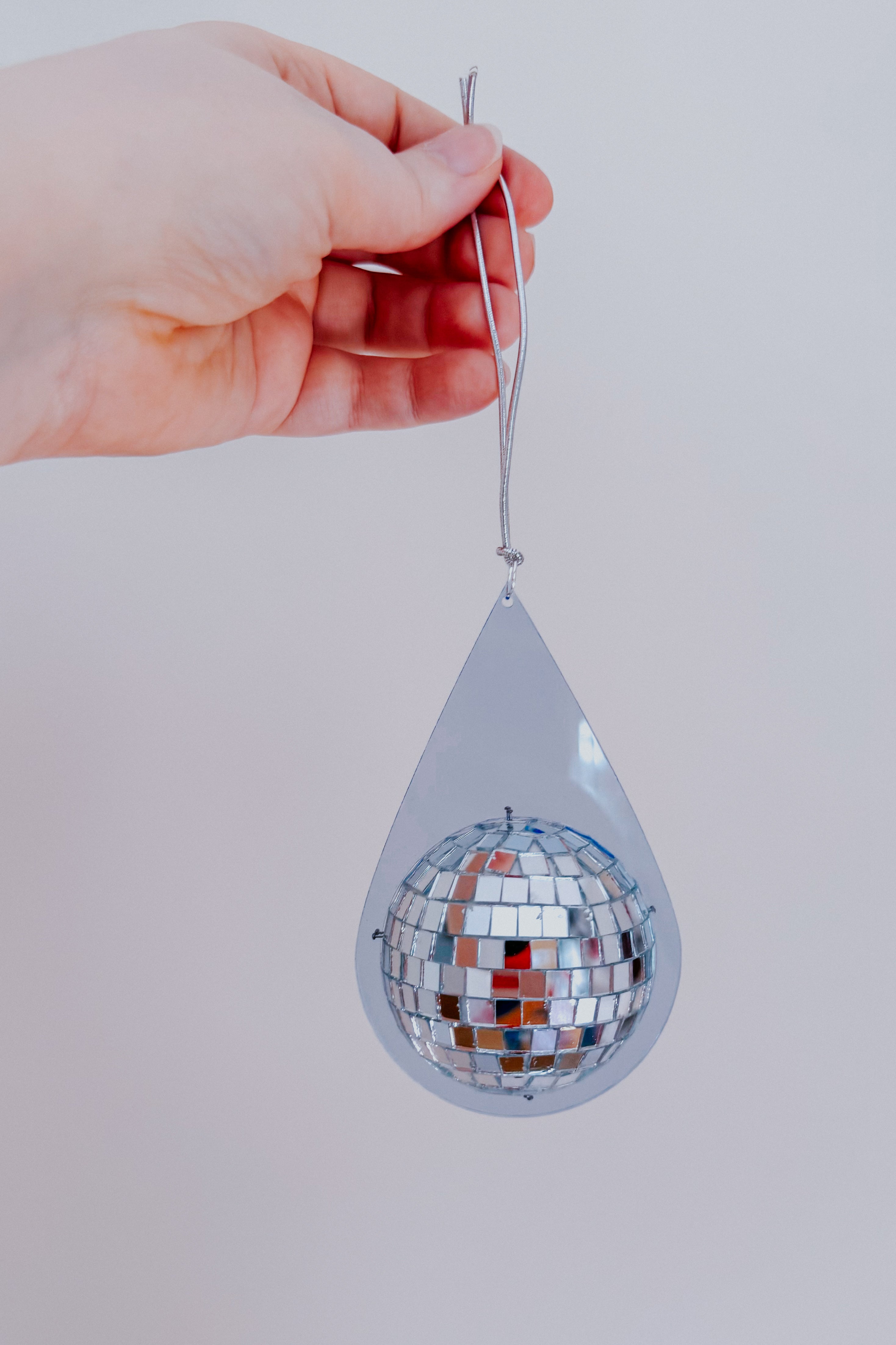 Hand holding light blue teardrop-shaped sun catcher with mirrored mini disco ball suspended on silver string against neutral background