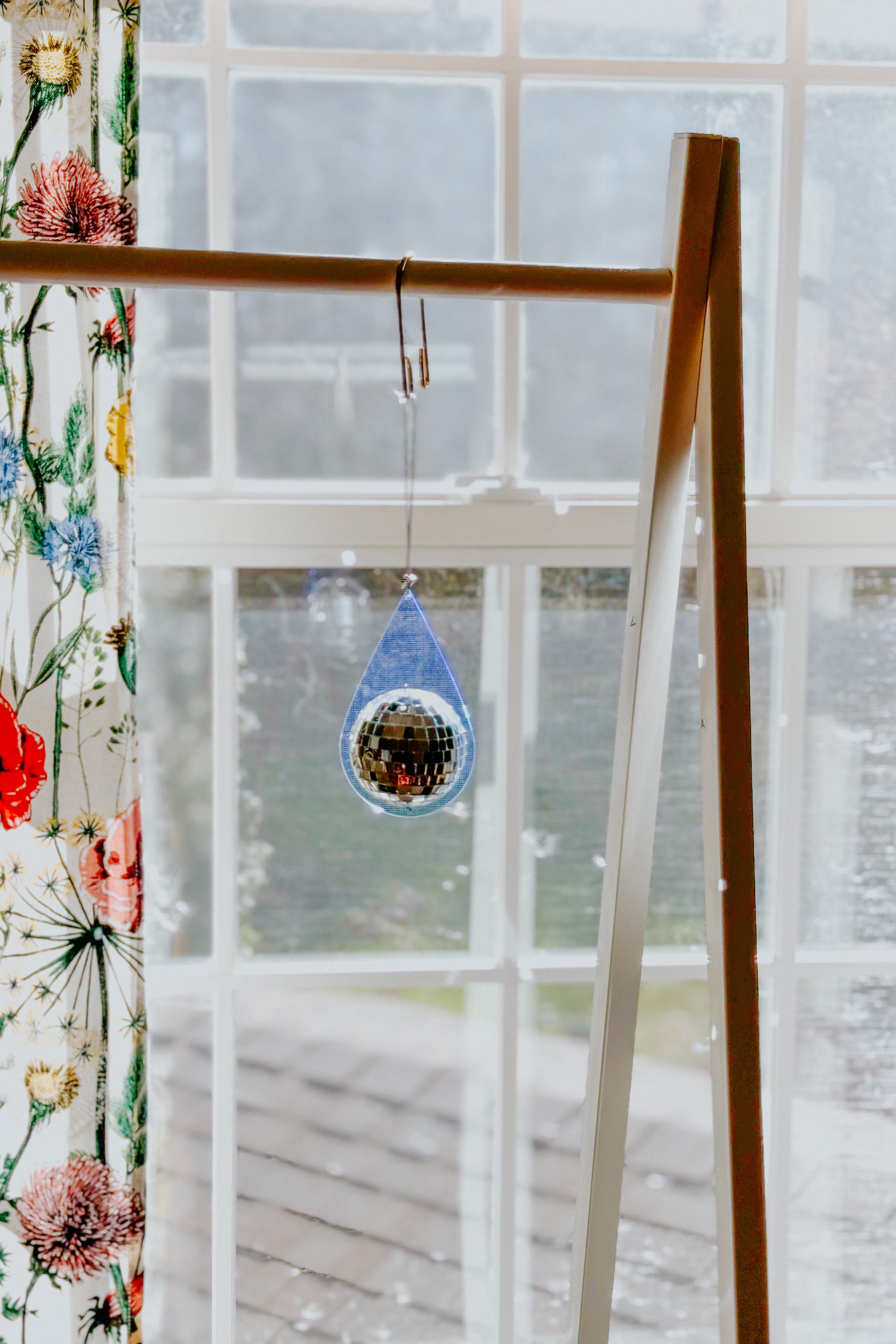 Light blue teardrop sun catcher with reflective mini disco ball hanging from wooden frame near window with floral curtain