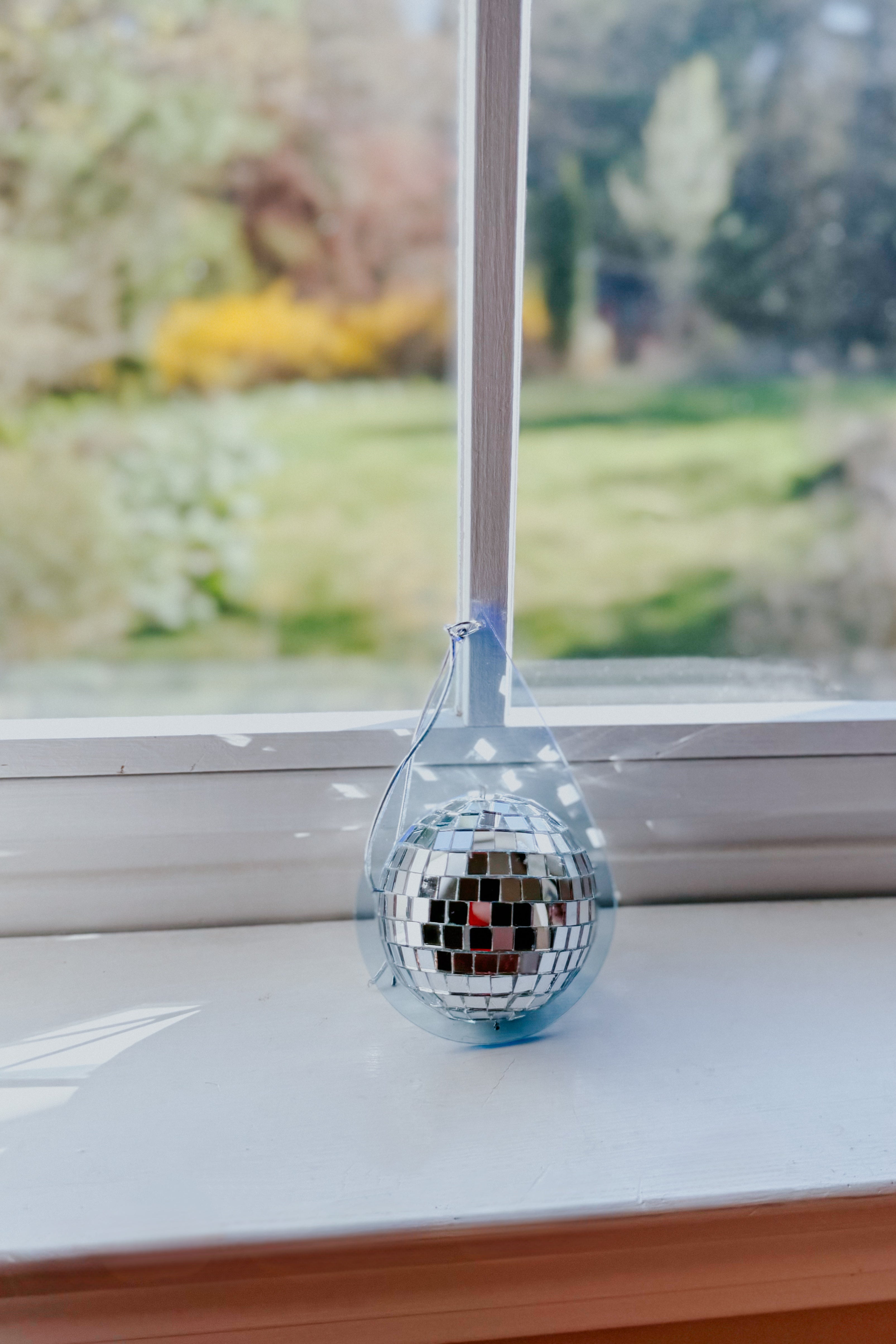 Light blue teardrop sun catcher with hanging reflective mini disco ball resting on white windowsill with garden view beyond