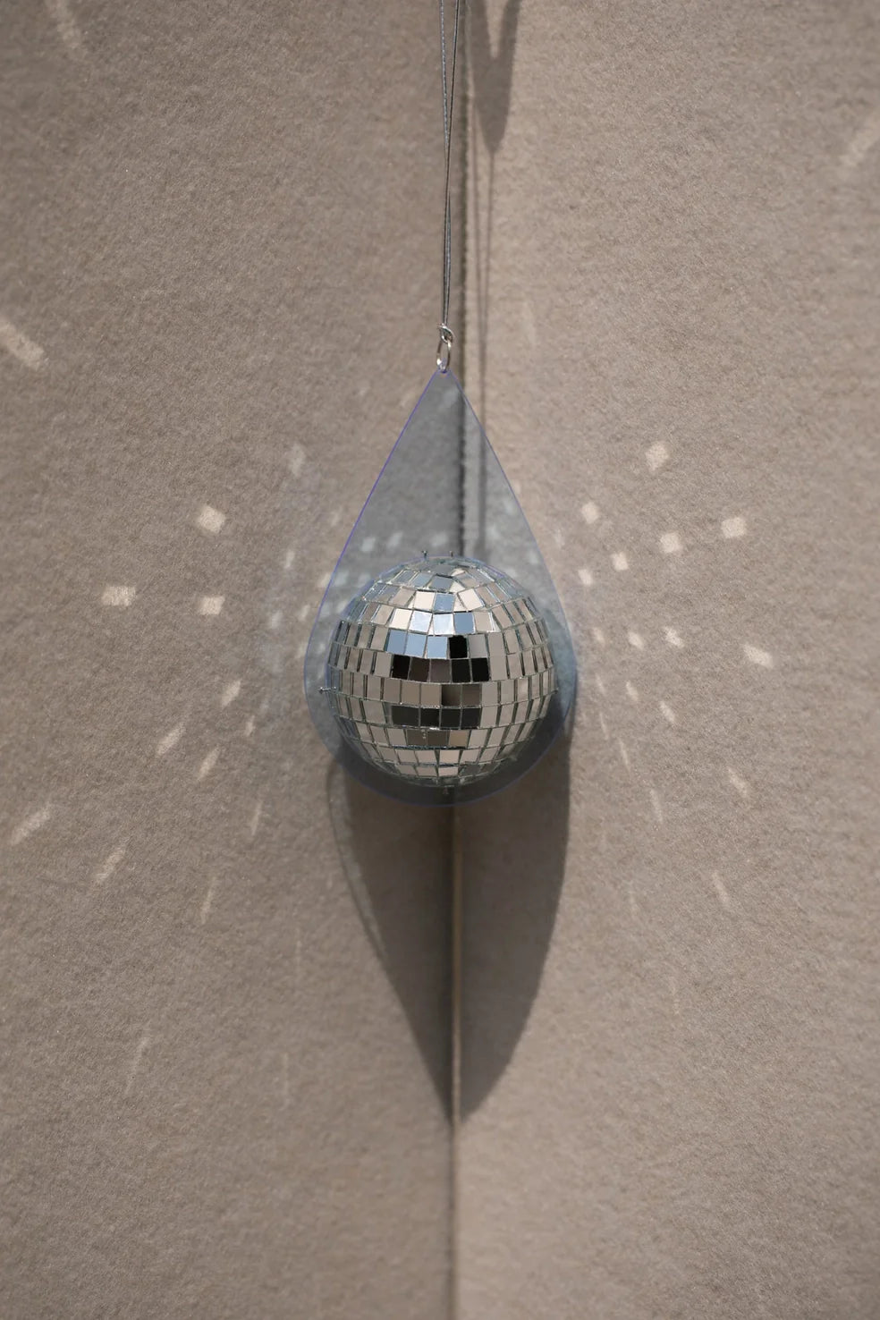 Light blue teardrop-shaped sun catcher with a hanging silver mini disco ball casting reflective light spots on beige wall