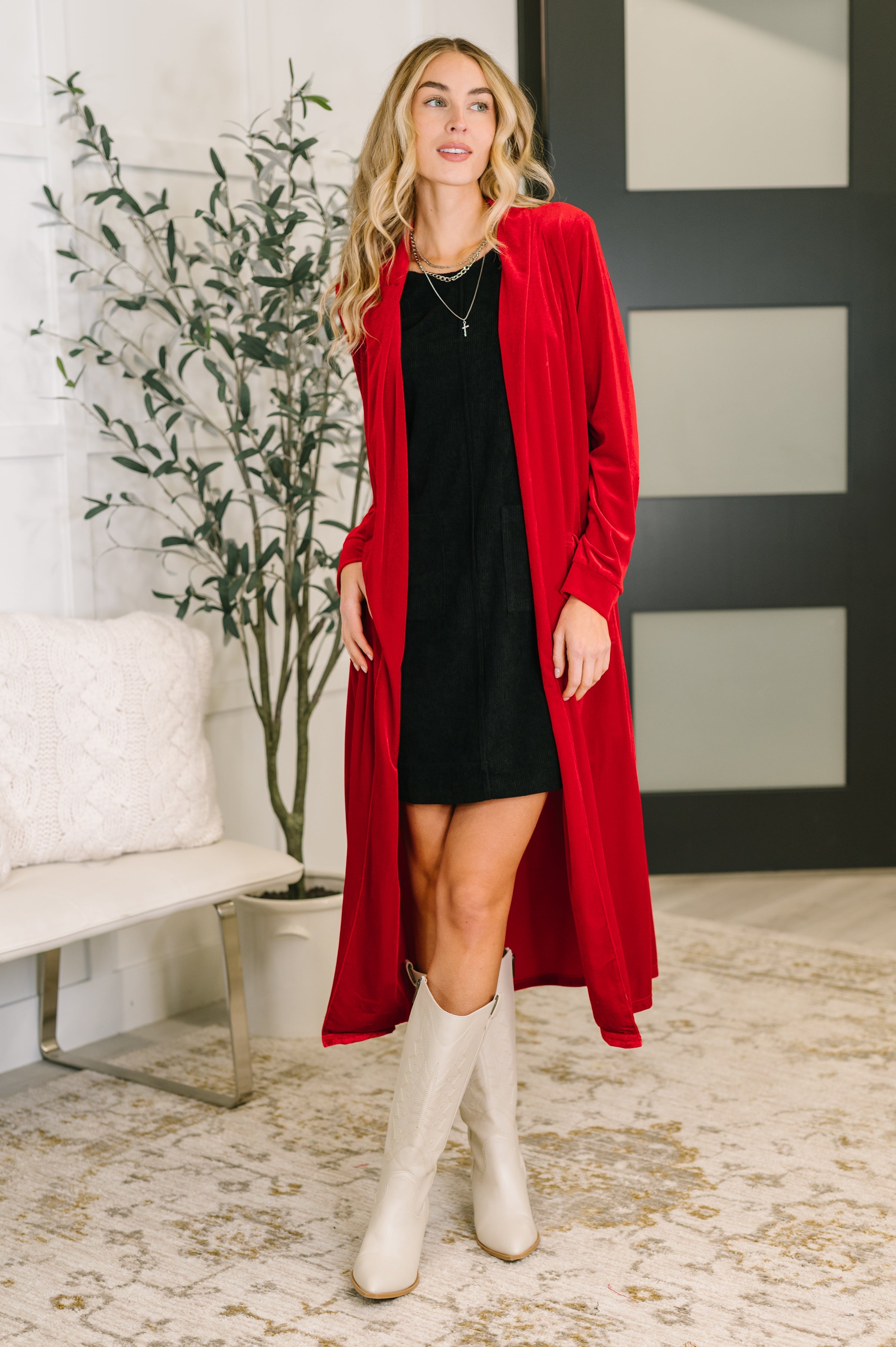 Woman wearing a red velvet cardigan, a black dress, and white boots in an indoor setting.