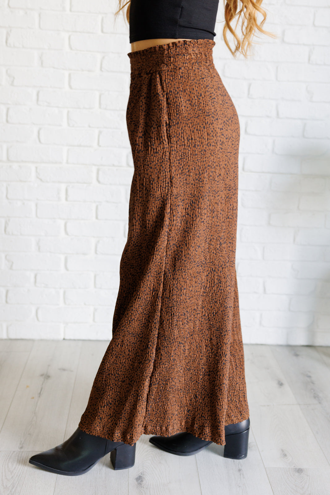 Side view of brown Harmony high rise wide leg pants with textured fabric worn with black cropped top and black heeled boots indoors