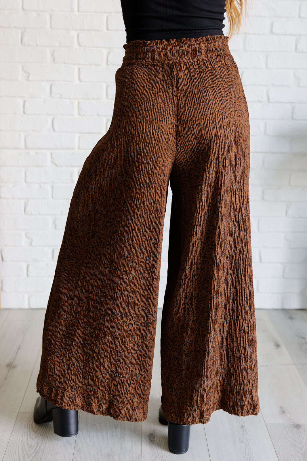 Side view of brown Harmony high rise wide leg pants with textured fabric worn with black top and boots indoors