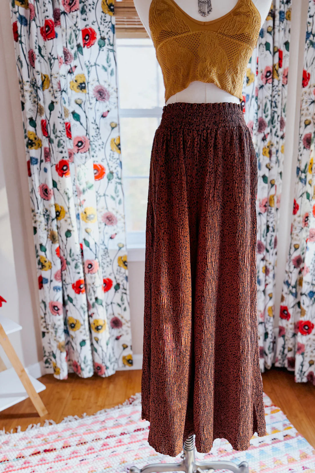 Brown high rise wide leg pants with textured fabric displayed on mannequin in bright room with floral curtains background
