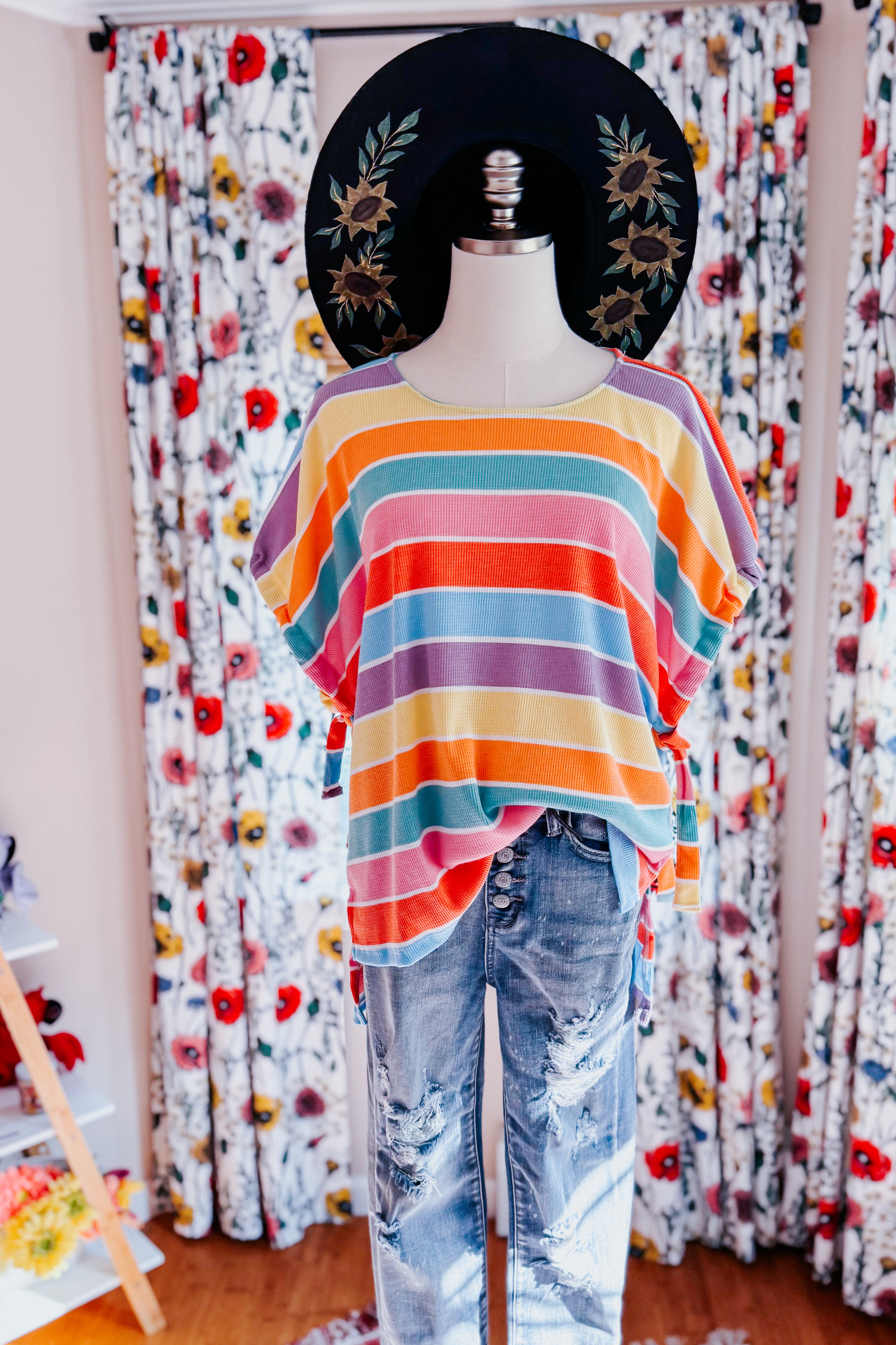 Multicolor striped dolman sleeve top paired with distressed denim jeans and black wide-brim hat on mannequin indoors