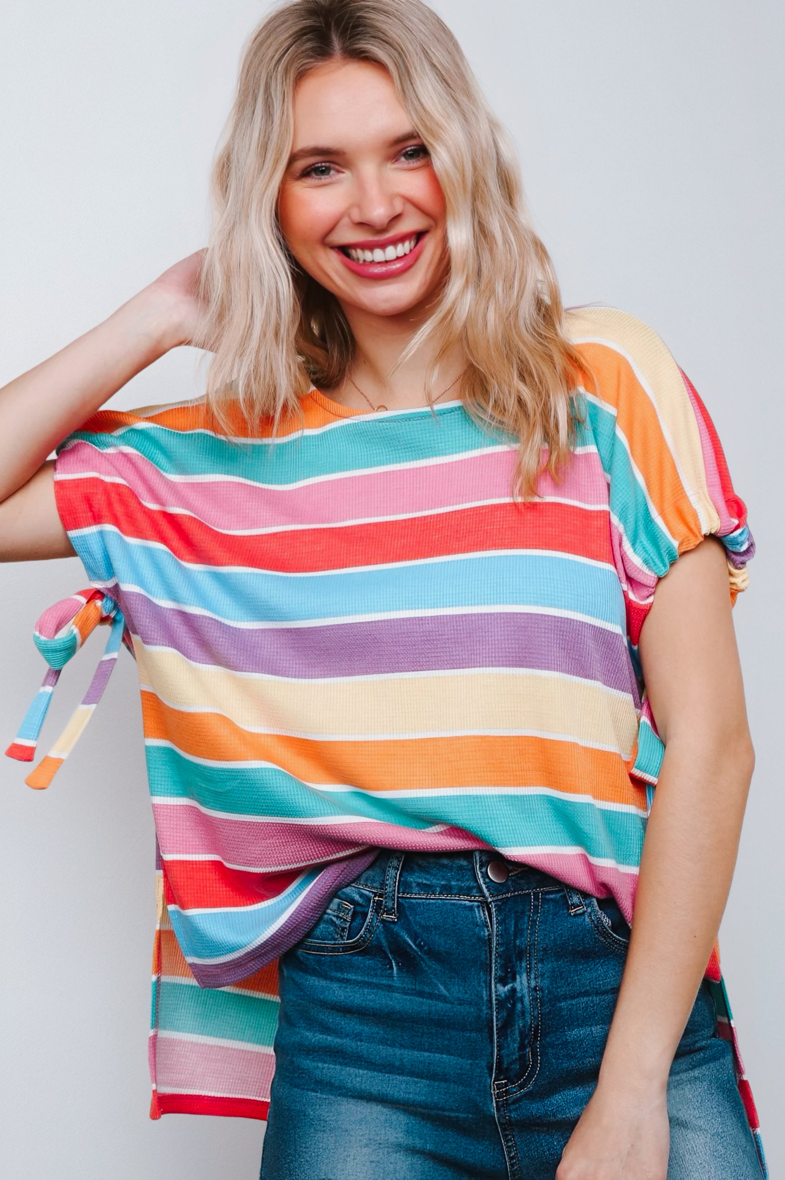 Woman smiling in multicolor striped dolman sleeve top with side tie detail paired with high-waisted denim jeans against white background