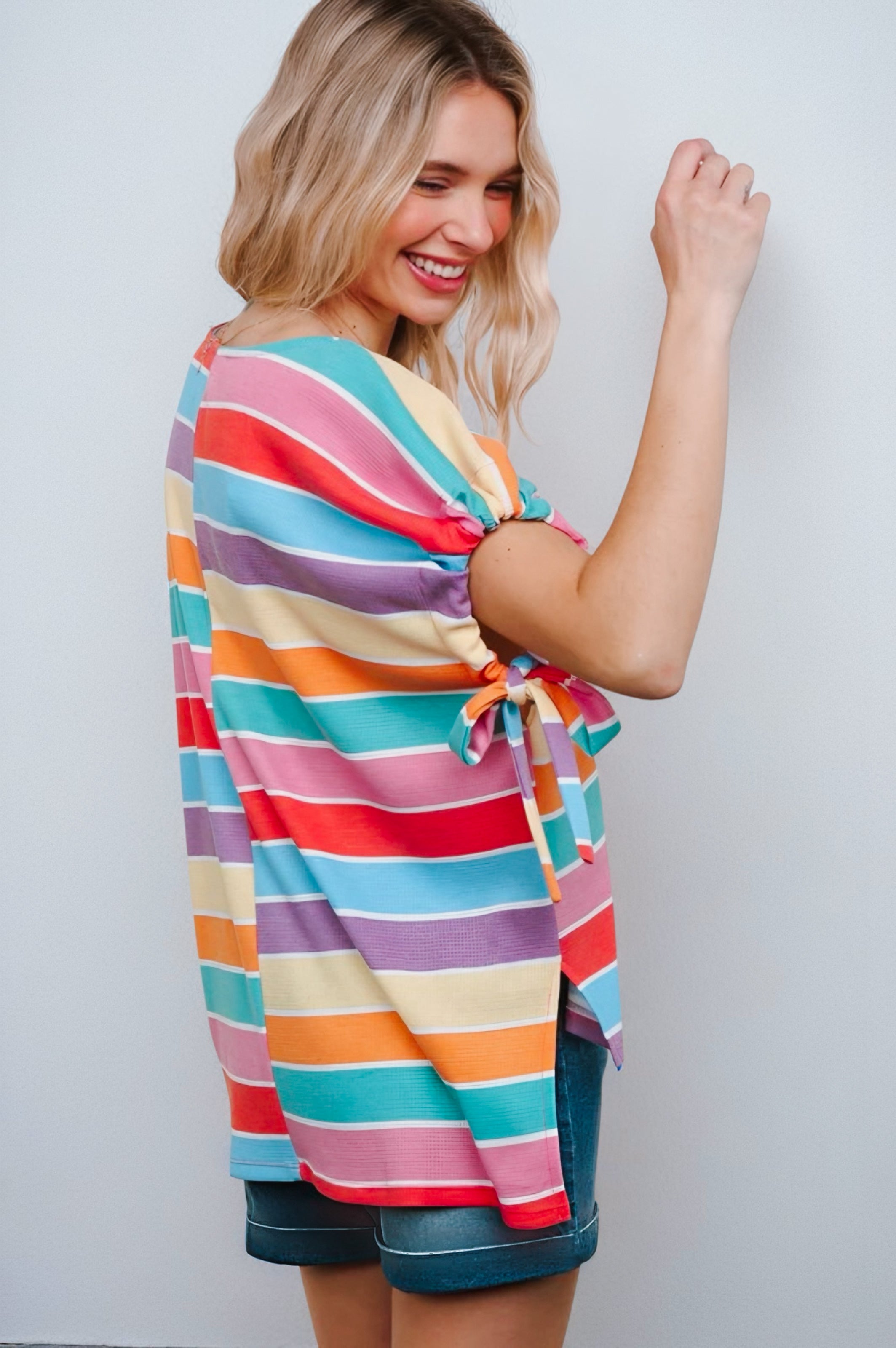Smiling woman wearing multicolor striped dolman sleeve top with side tie detail paired with denim shorts against neutral background