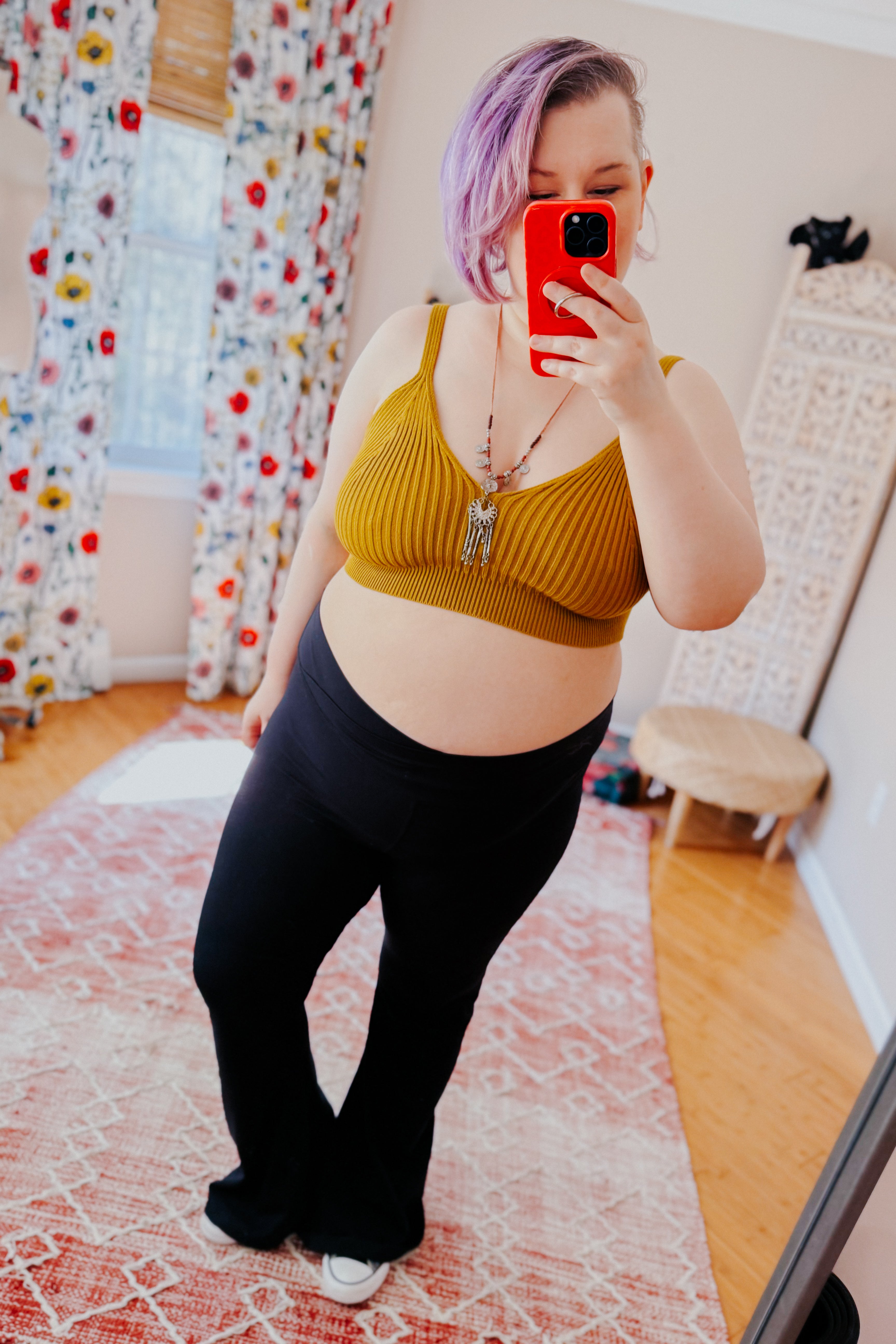 Woman with purple hair taking selfie wearing mustard rib knit brami bralette and black high-waisted pants indoors on patterned rug