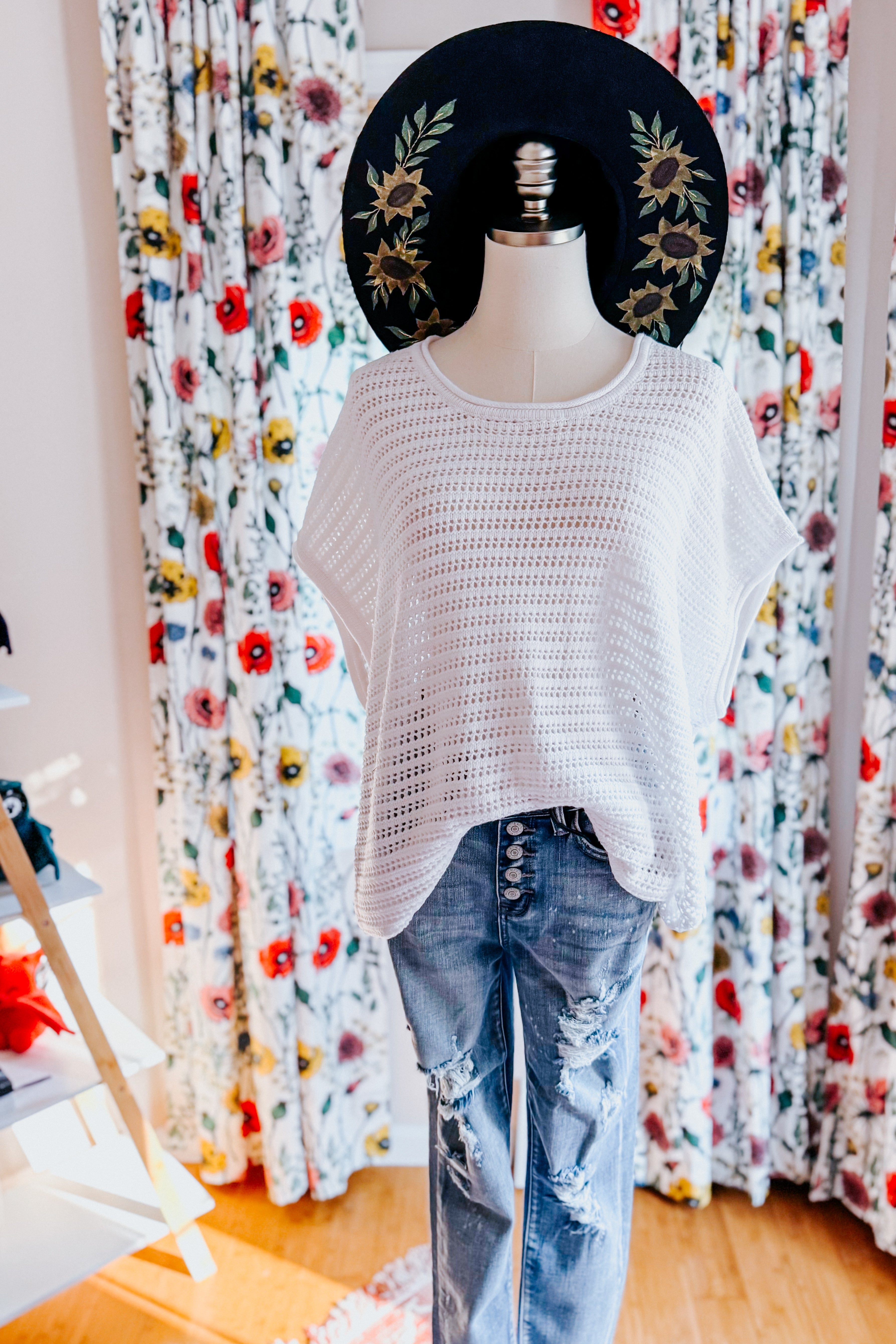 White open knit cap sleeve sweater styled loosely over distressed button-fly denim jeans on mannequin with floral curtains background