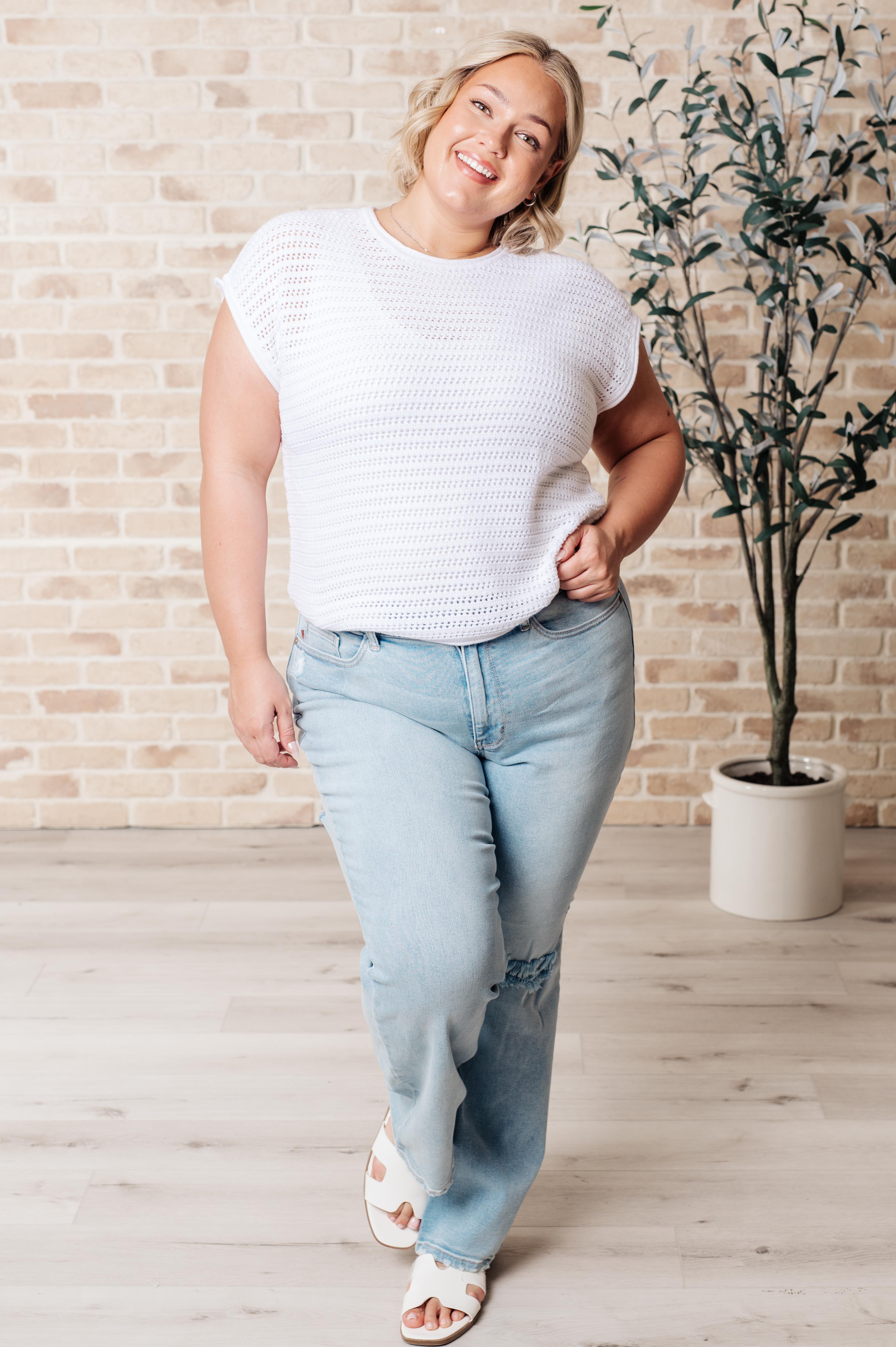Smiling woman wearing white open knit cap sleeve sweater paired with light wash ripped denim jeans indoors