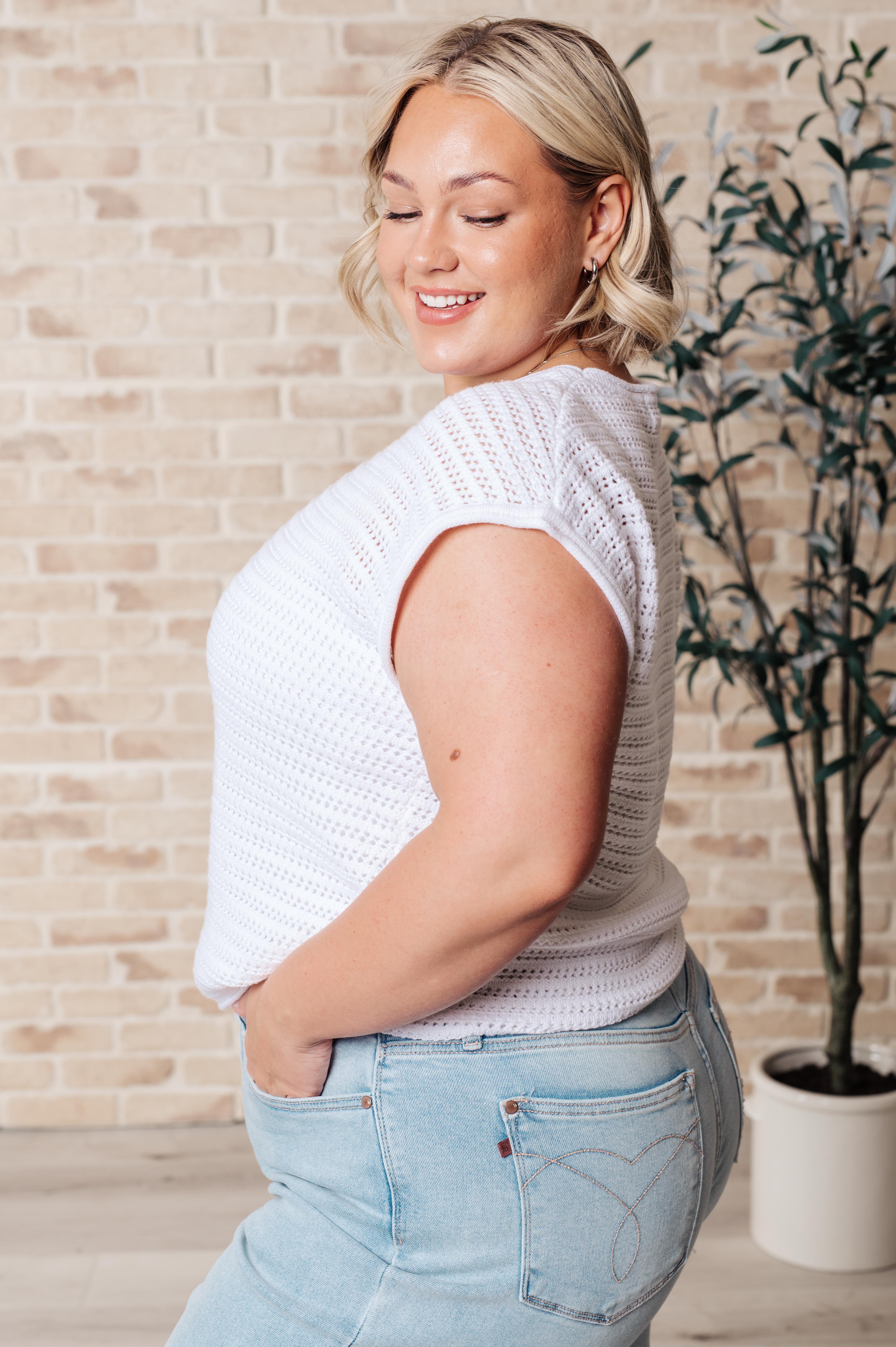 Woman wearing white open knit cap sleeve sweater paired with light blue denim jeans standing indoors