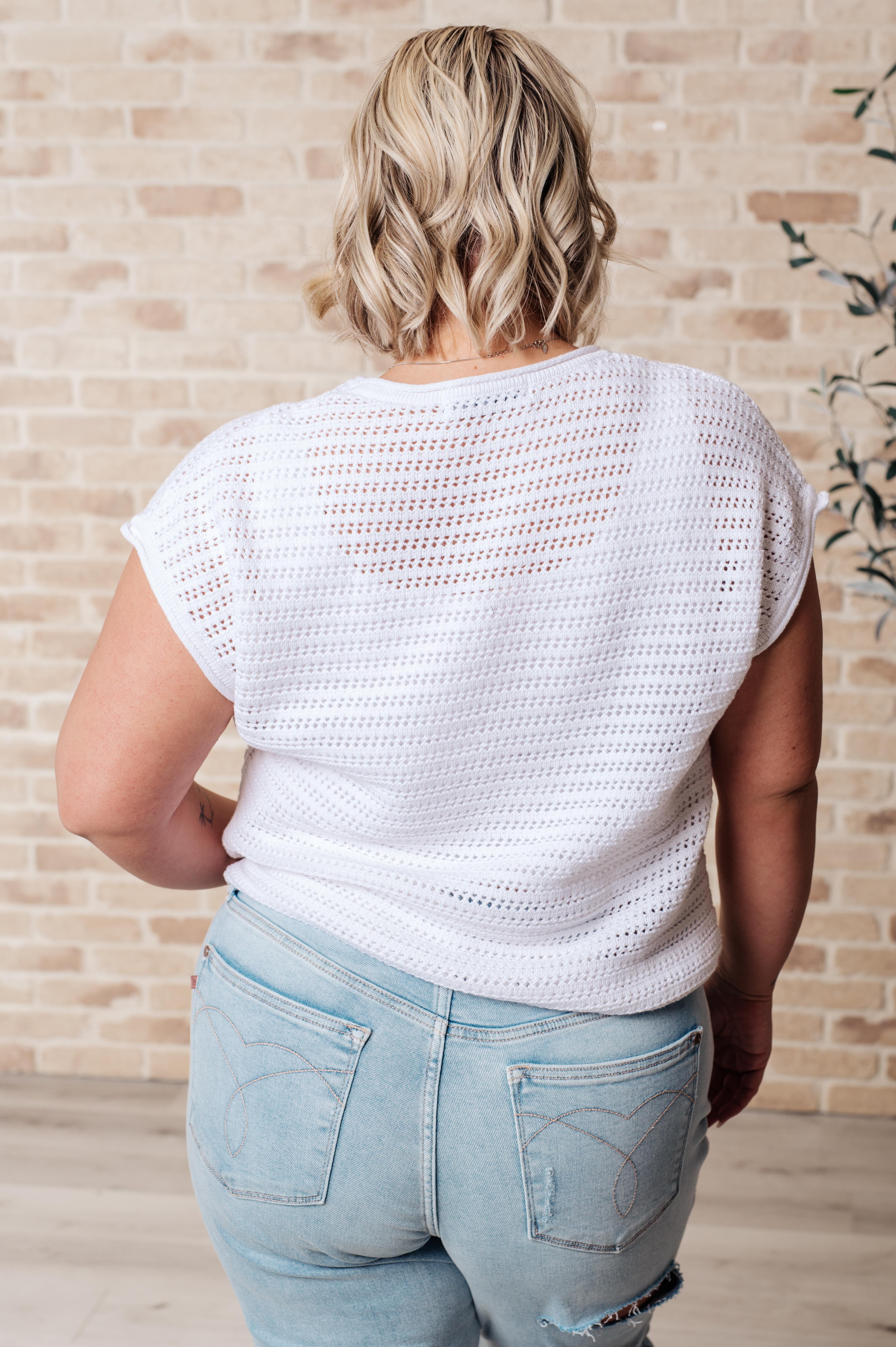 Back view of person wearing white open knit cap sleeve sweater paired with light wash denim jeans against brick wall