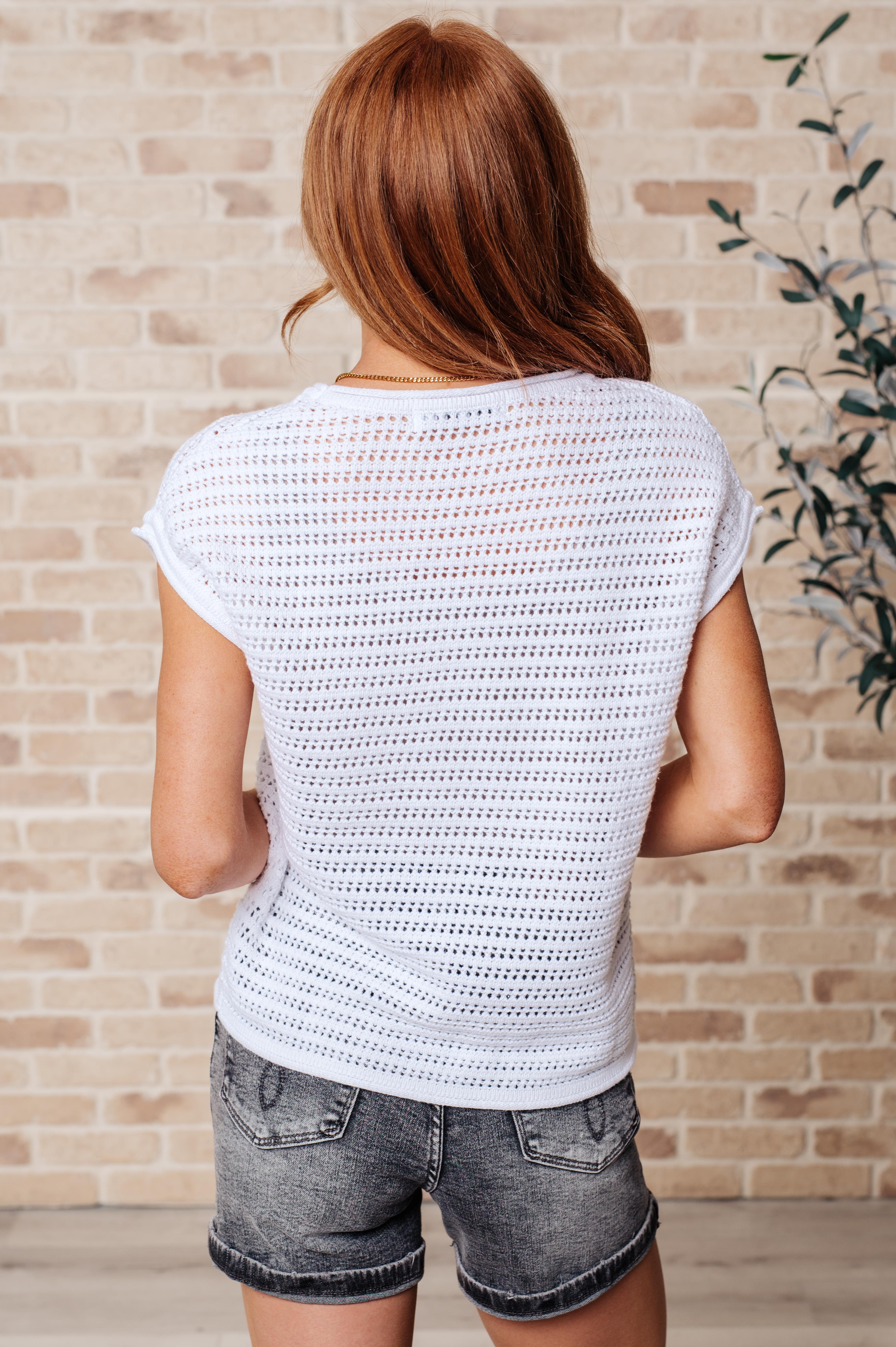 Back view of woman wearing white open knit cap sleeve sweater paired with dark denim shorts in casual setting