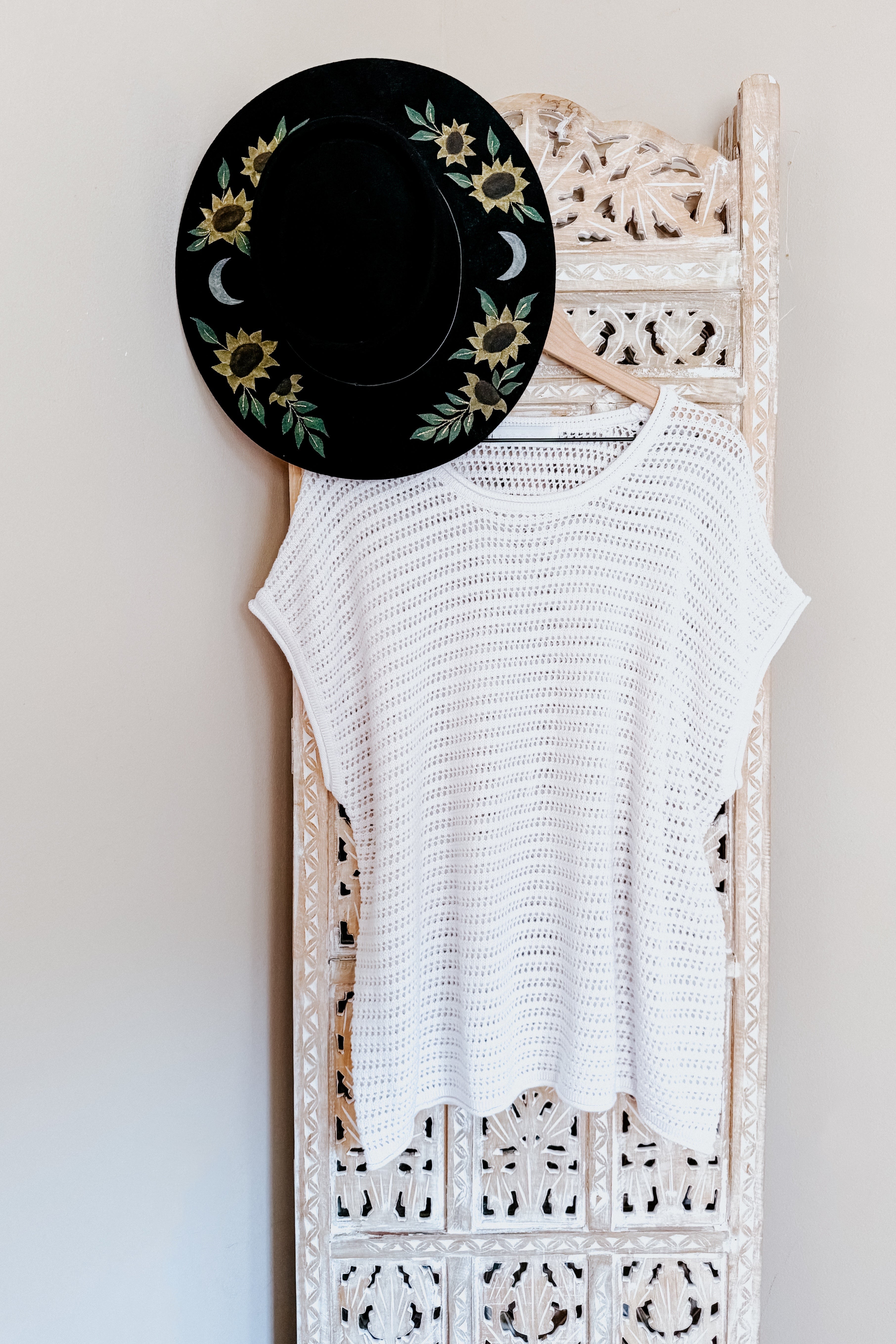 White open knit cap sleeve sweater hanging on ornate carved wooden screen with black wide-brimmed floral hat above