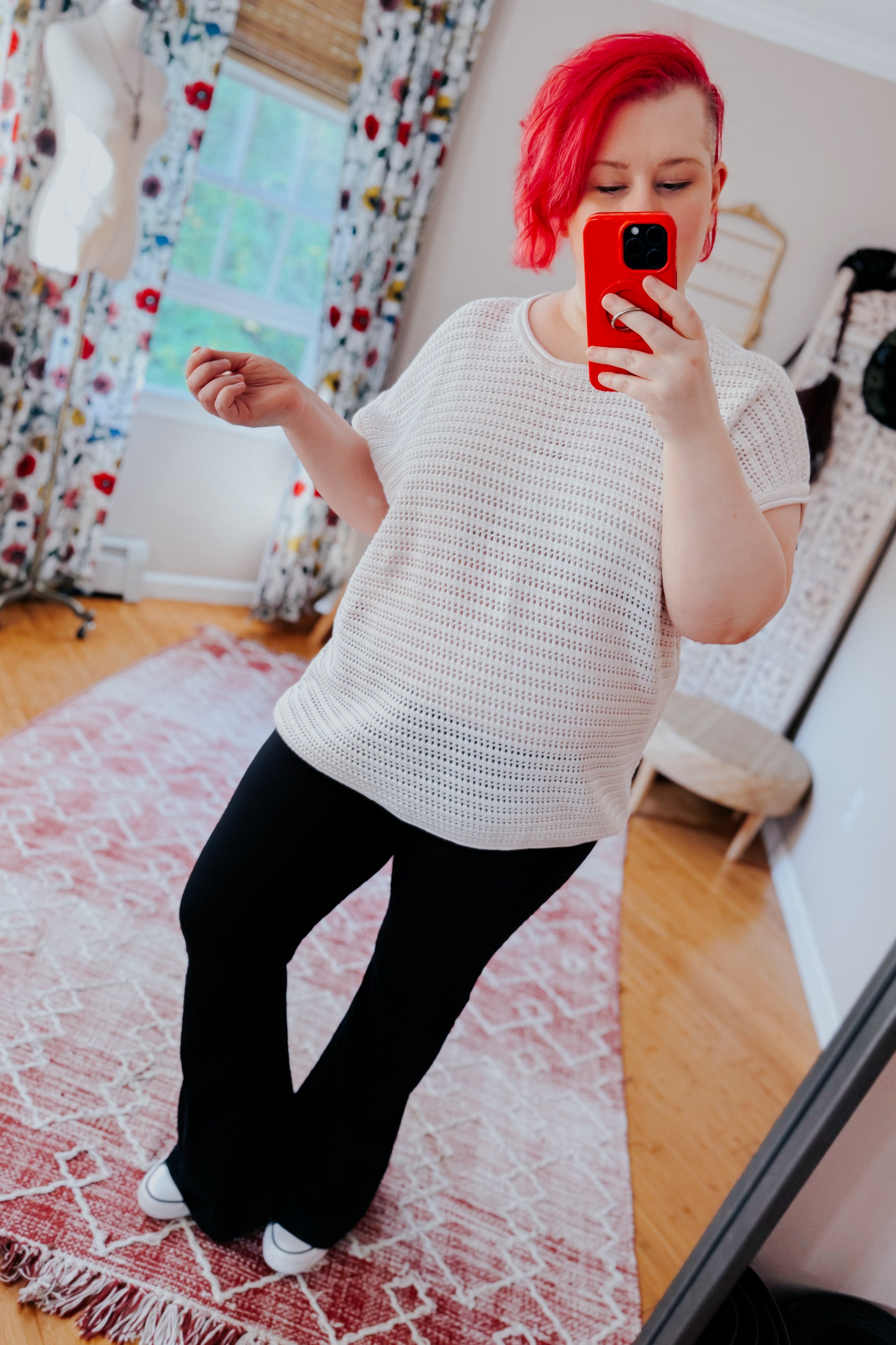 Woman with red hair wearing white open knit cap sleeve sweater and black pants taking selfie in bright room with patterned curtains