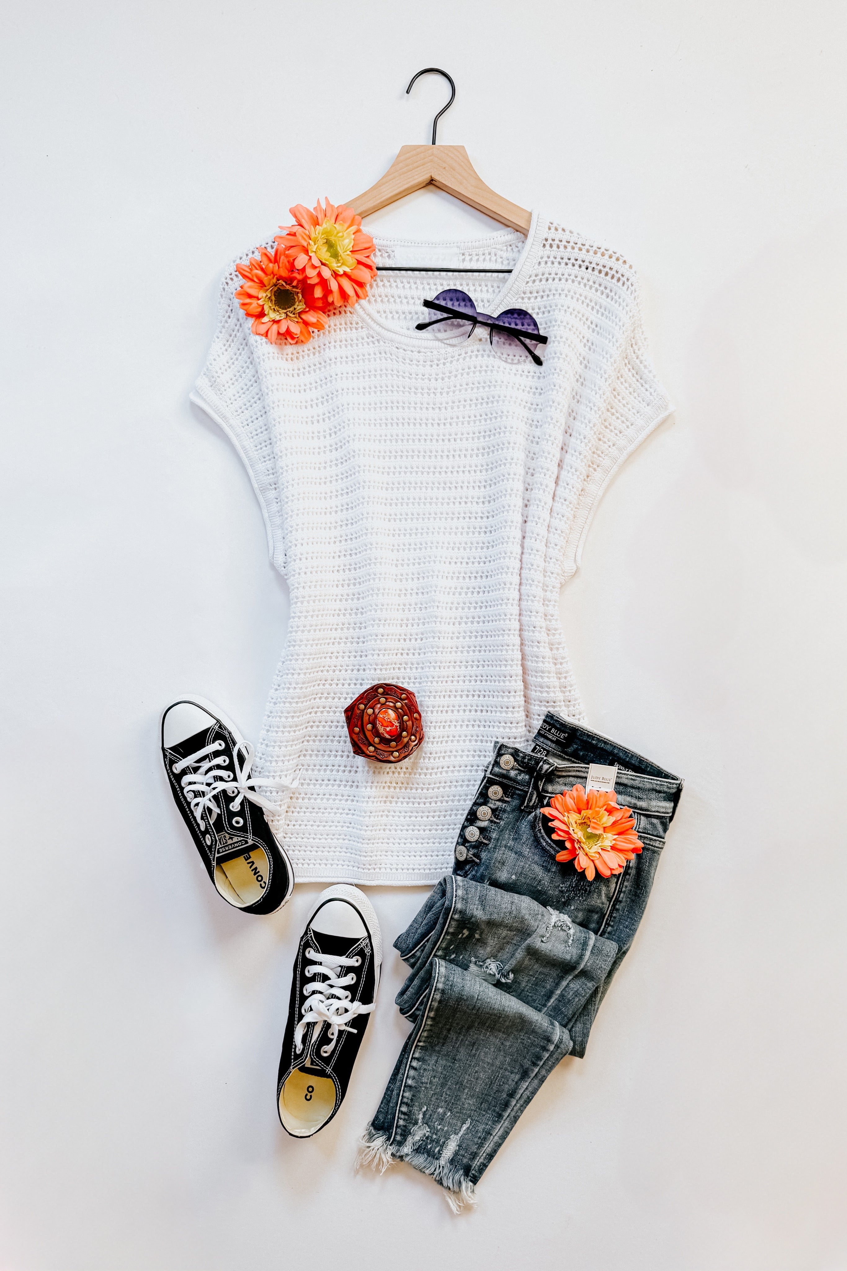 White open knit cap sleeve sweater styled with distressed denim jeans, black sneakers, sunglasses, and orange floral accents on white background