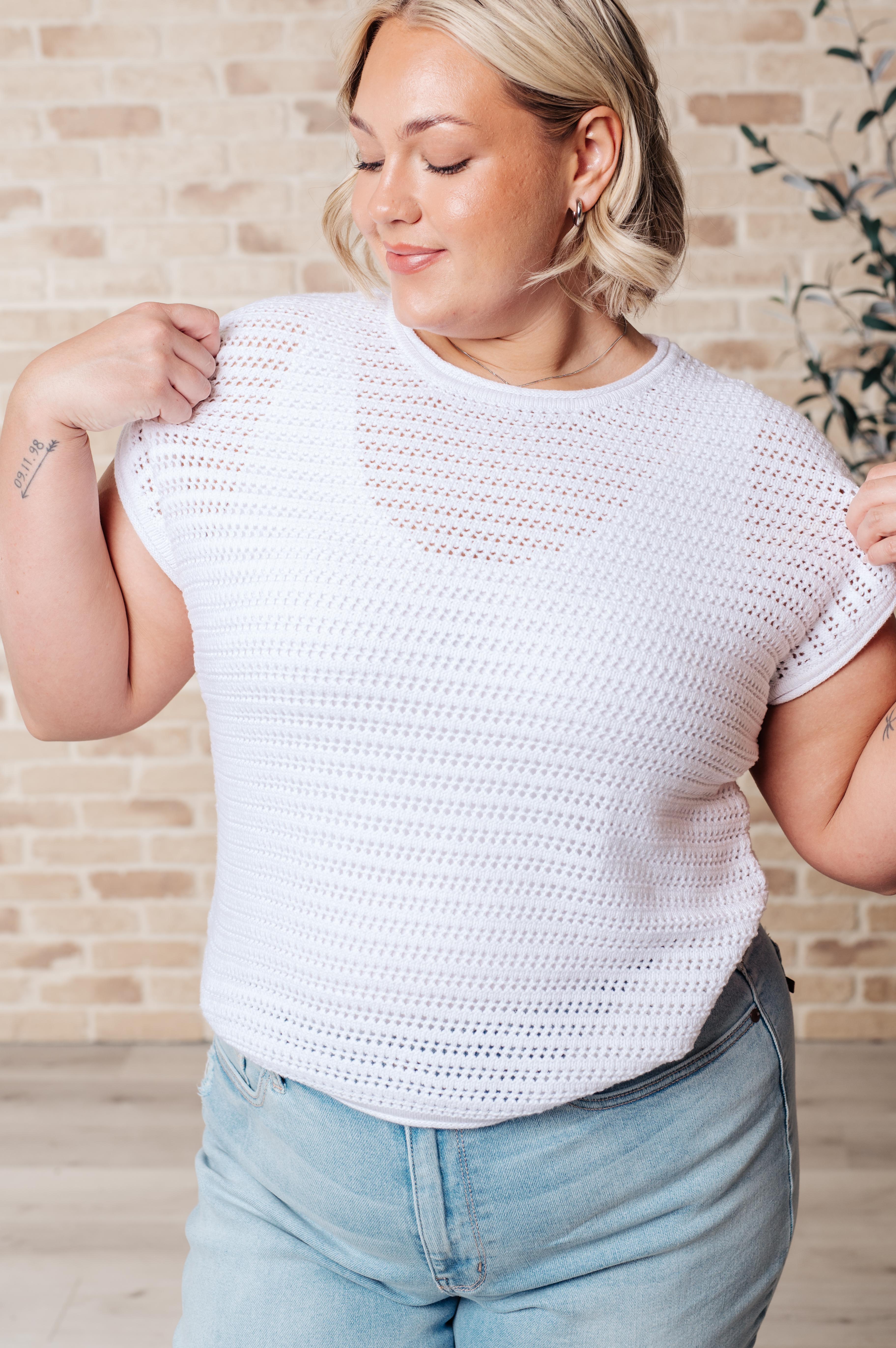 Smiling woman adjusting white open knit cap sleeve sweater paired with light blue denim jeans indoors with brick background