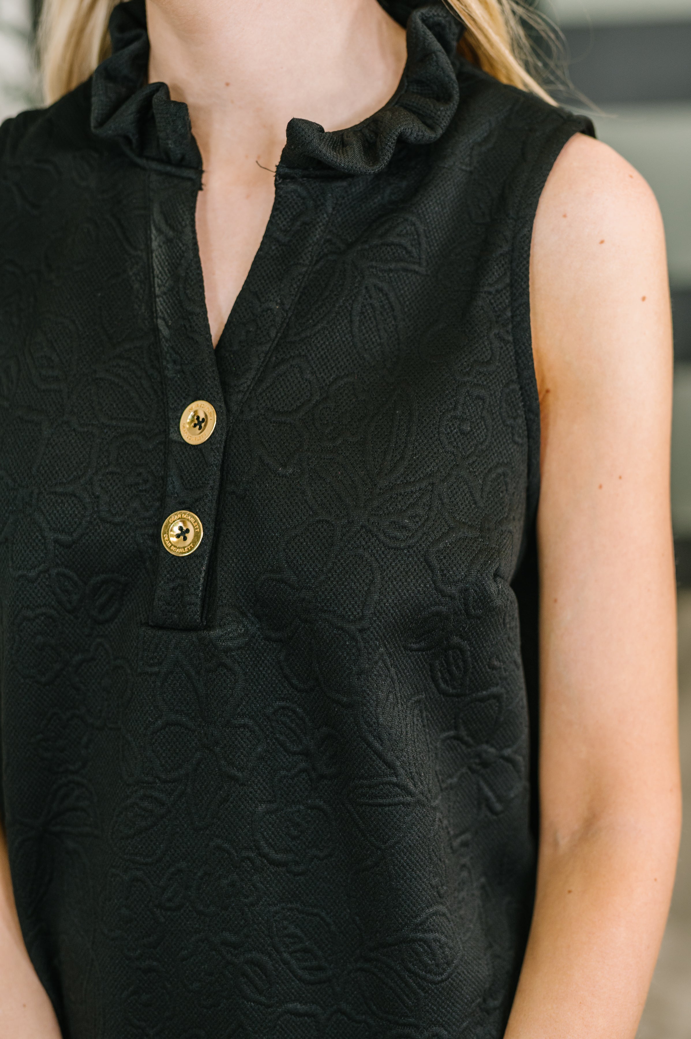 Close-up of sleeveless black embossed dress with floral pattern, ruffled high neck, and gold button accents on chest