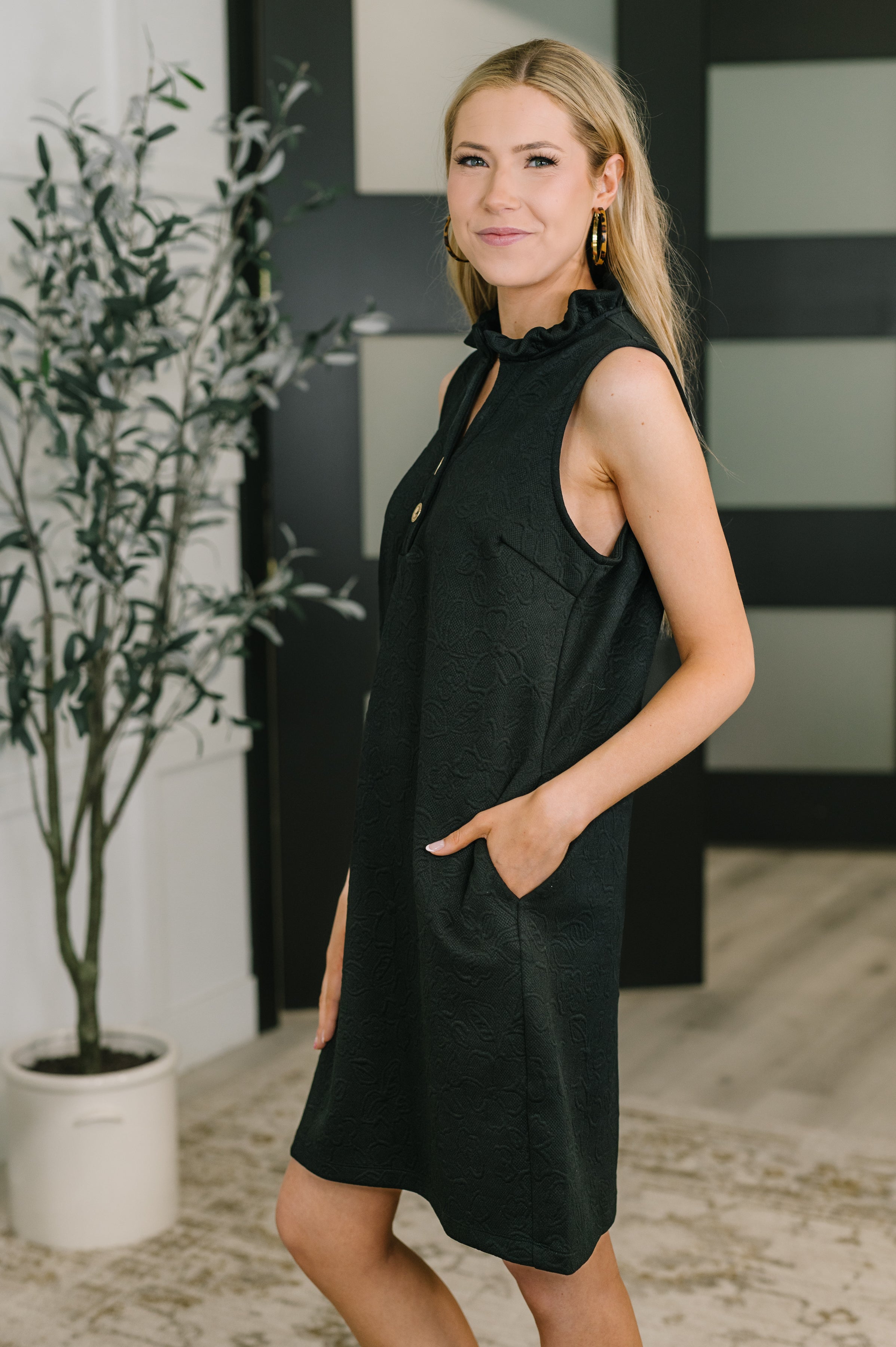 Woman in sleeveless black embossed dress with ruffled high neck and pockets standing indoors with hand in pocket