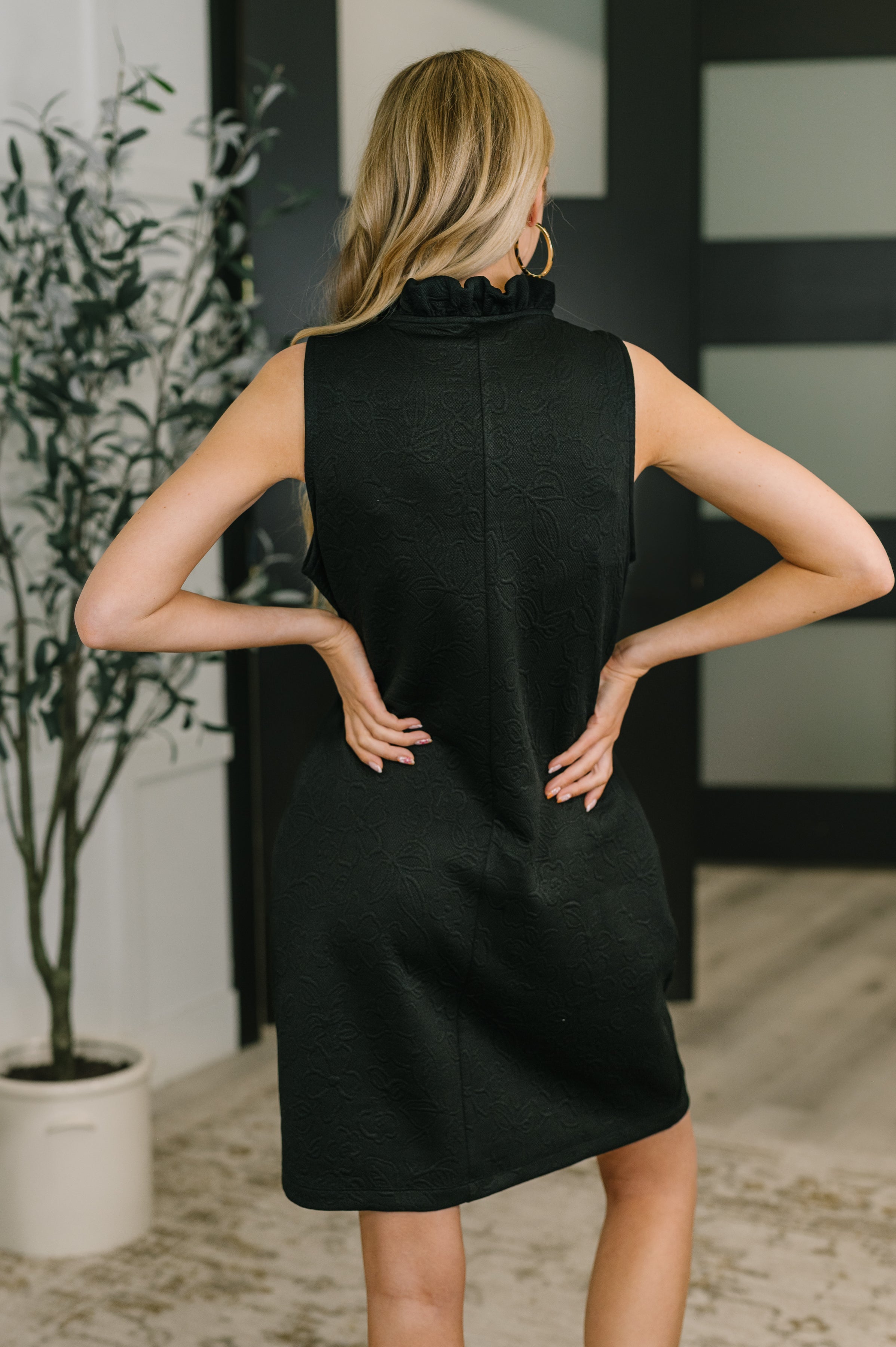 Back view of woman wearing sleeveless black embossed dress with ruffled high neck and hands on hips indoors