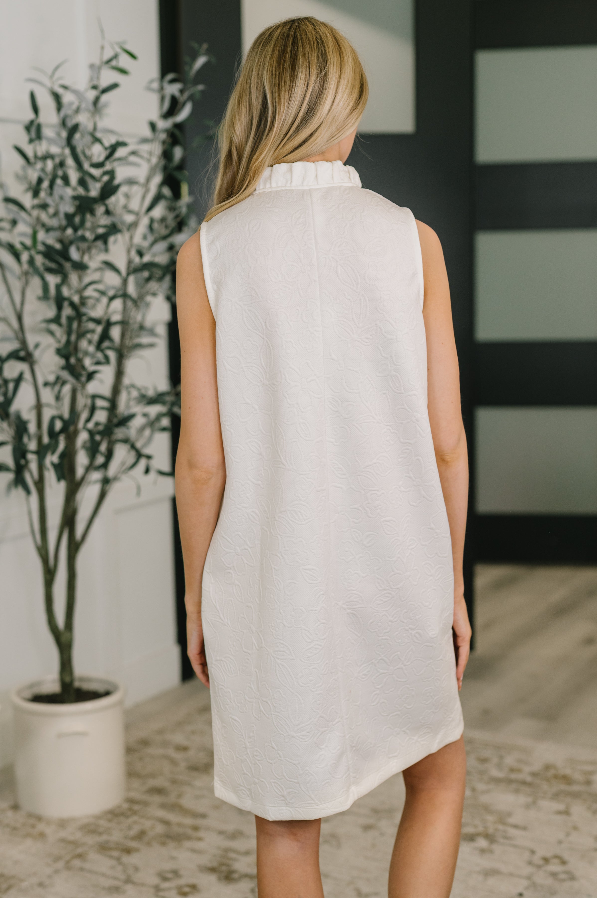 Back view of woman wearing off-white sleeveless dress with embossed texture and ruffled high neckline indoors