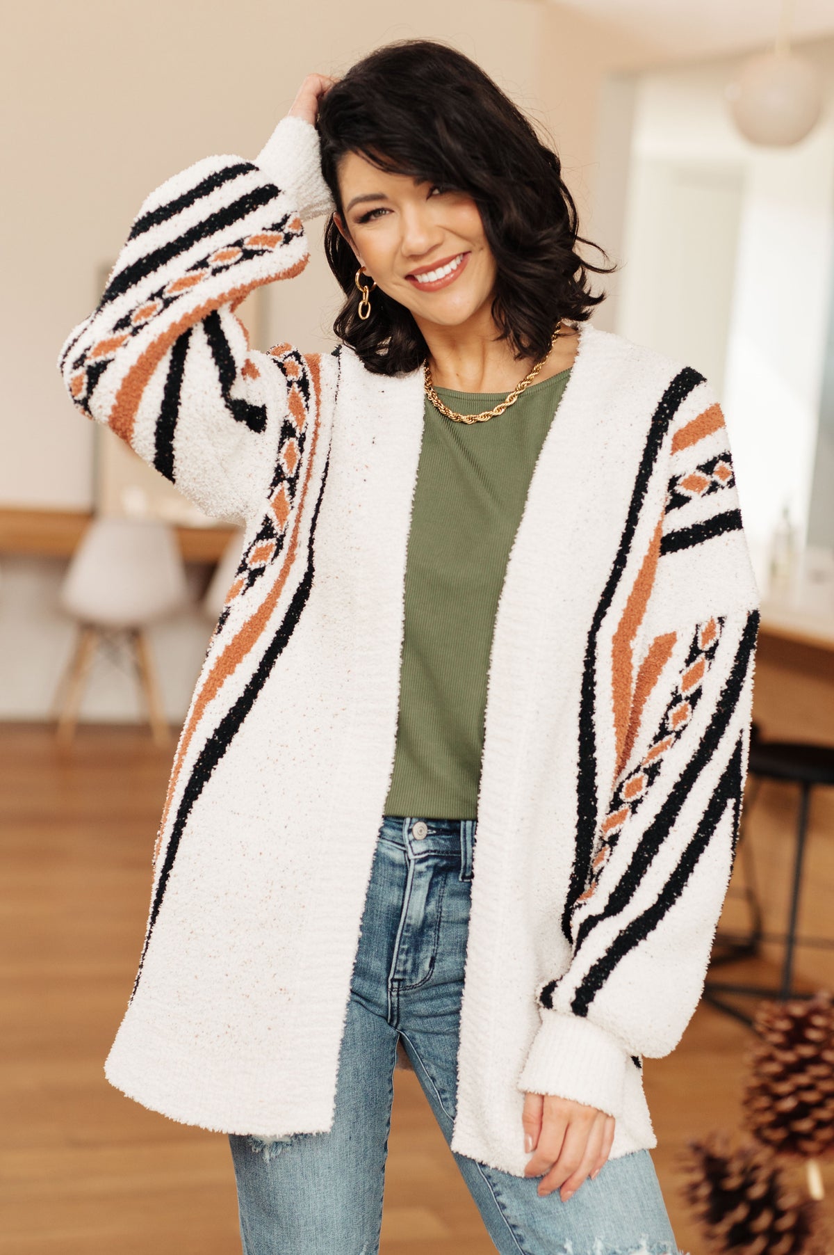 Woman smiling in a cream fuzzy open front cardigan with bold black and orange stripes styled with a green top and jeans indoors