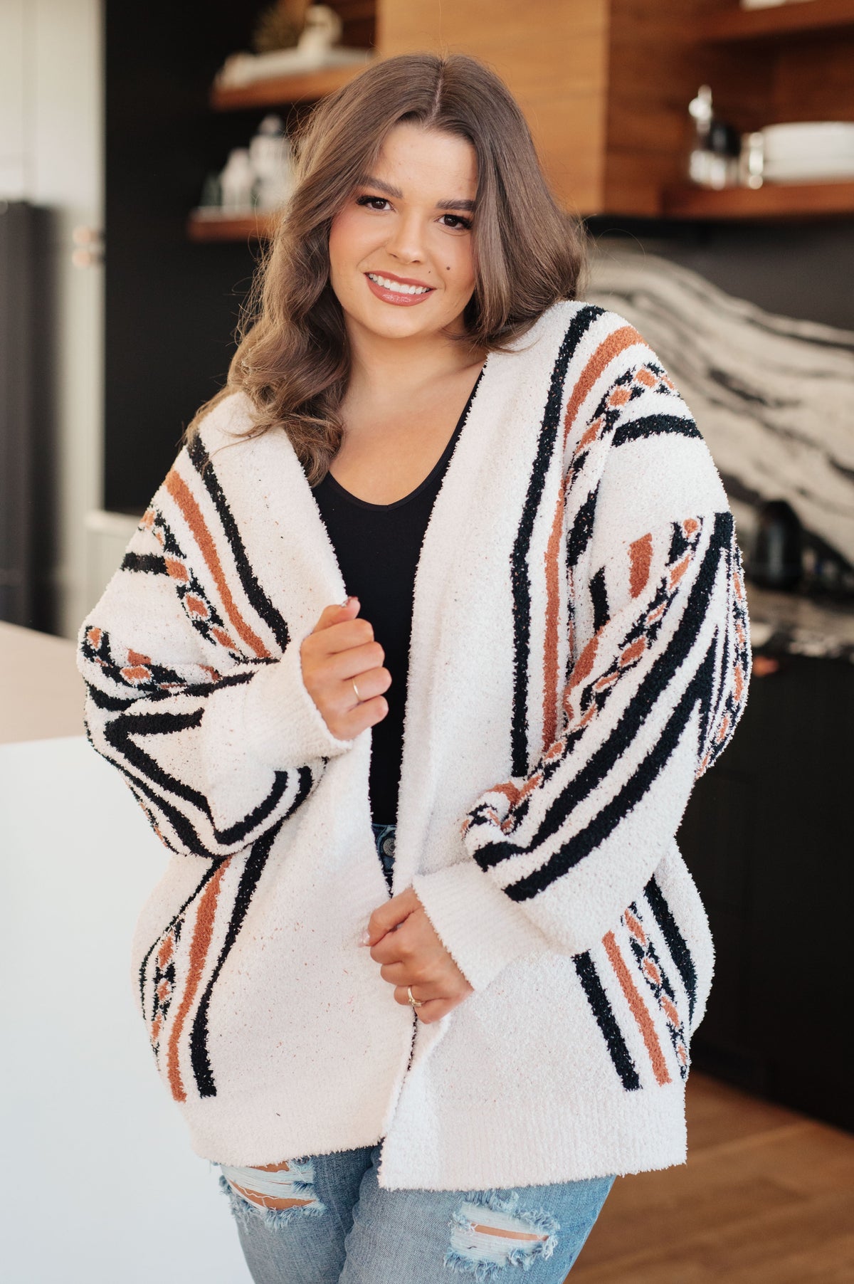Smiling woman wearing a cream fuzzy open front cardigan with black and rust patterns styled over a black top and ripped jeans indoors
