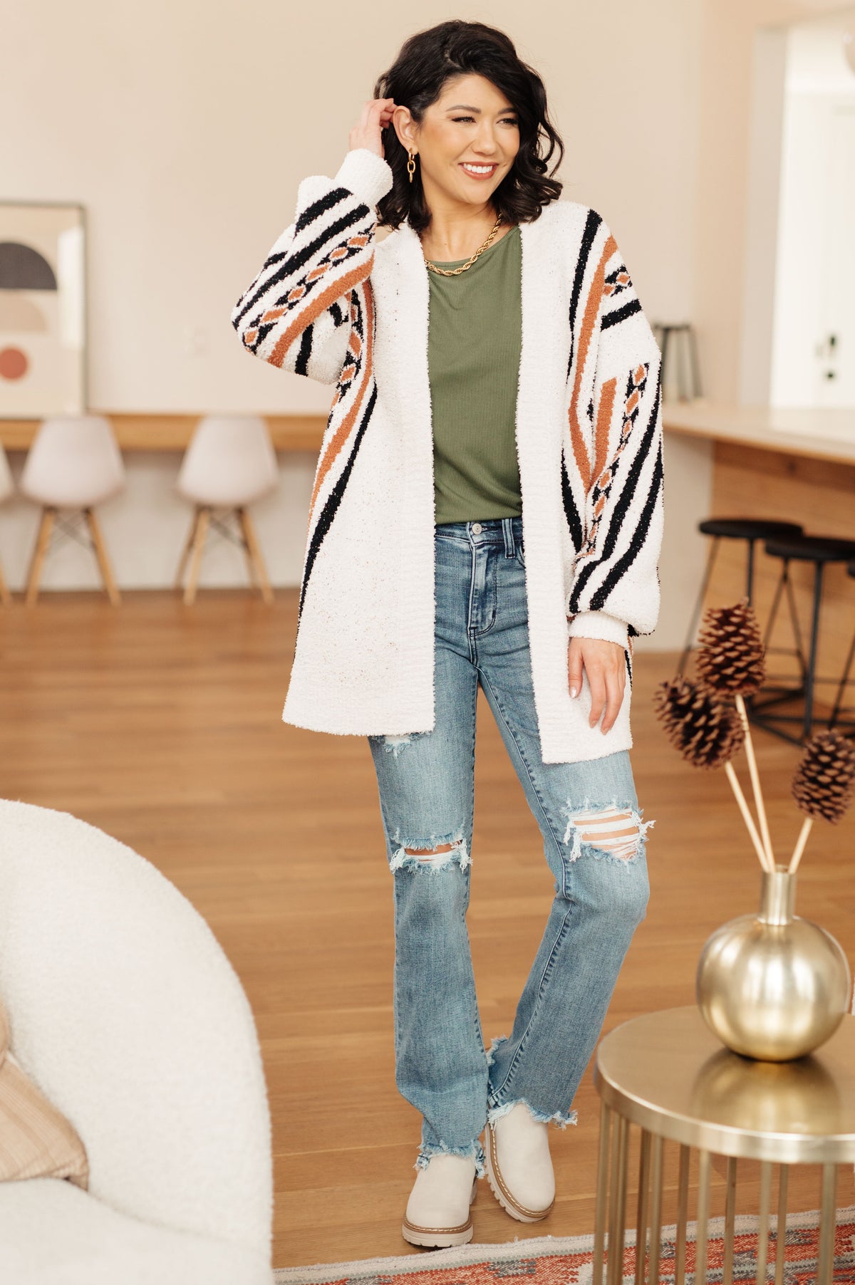 Woman wearing a cream fuzzy knit cardigan with bold striped patterns over an olive top and distressed jeans in a modern home