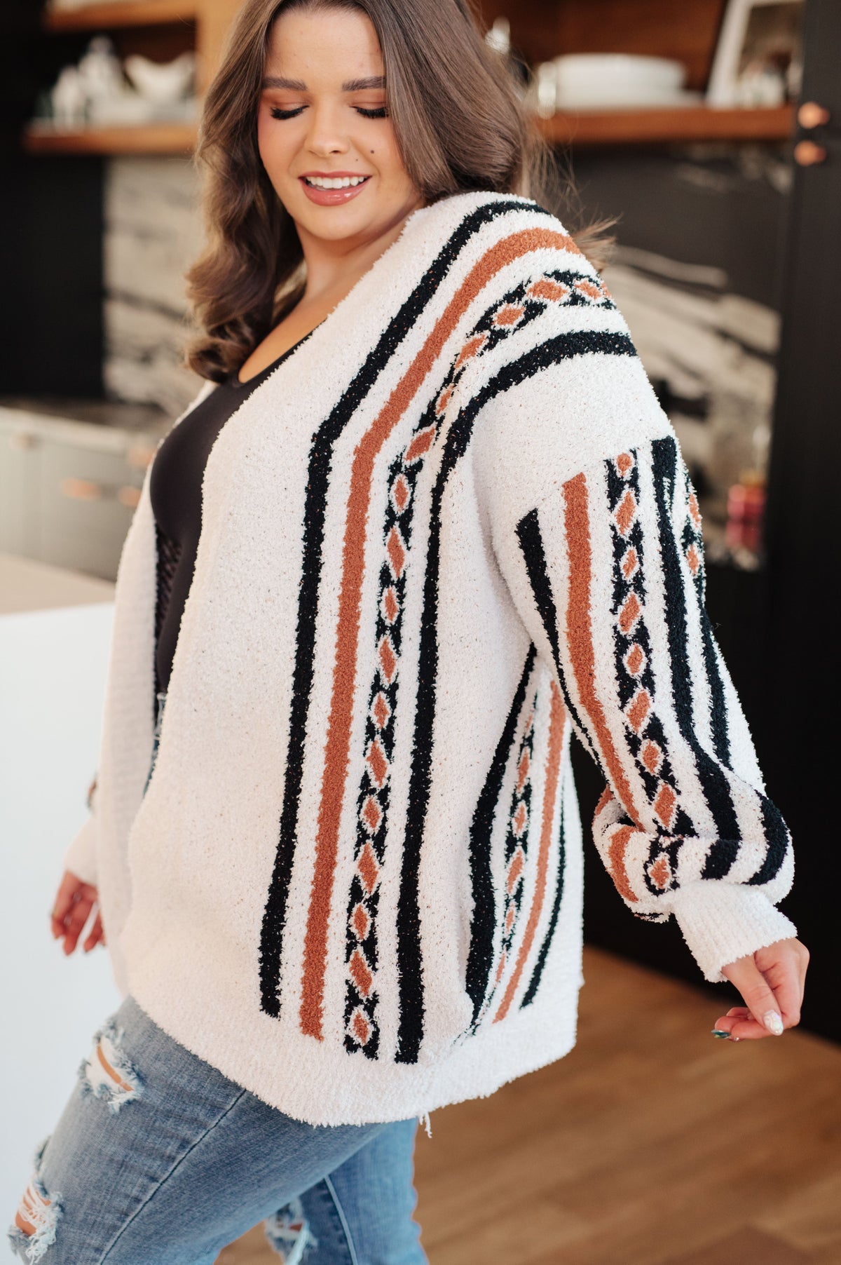 Smiling woman wearing a cream fuzzy knit cardigan with black and rust geometric stripes over a black top and jeans indoors