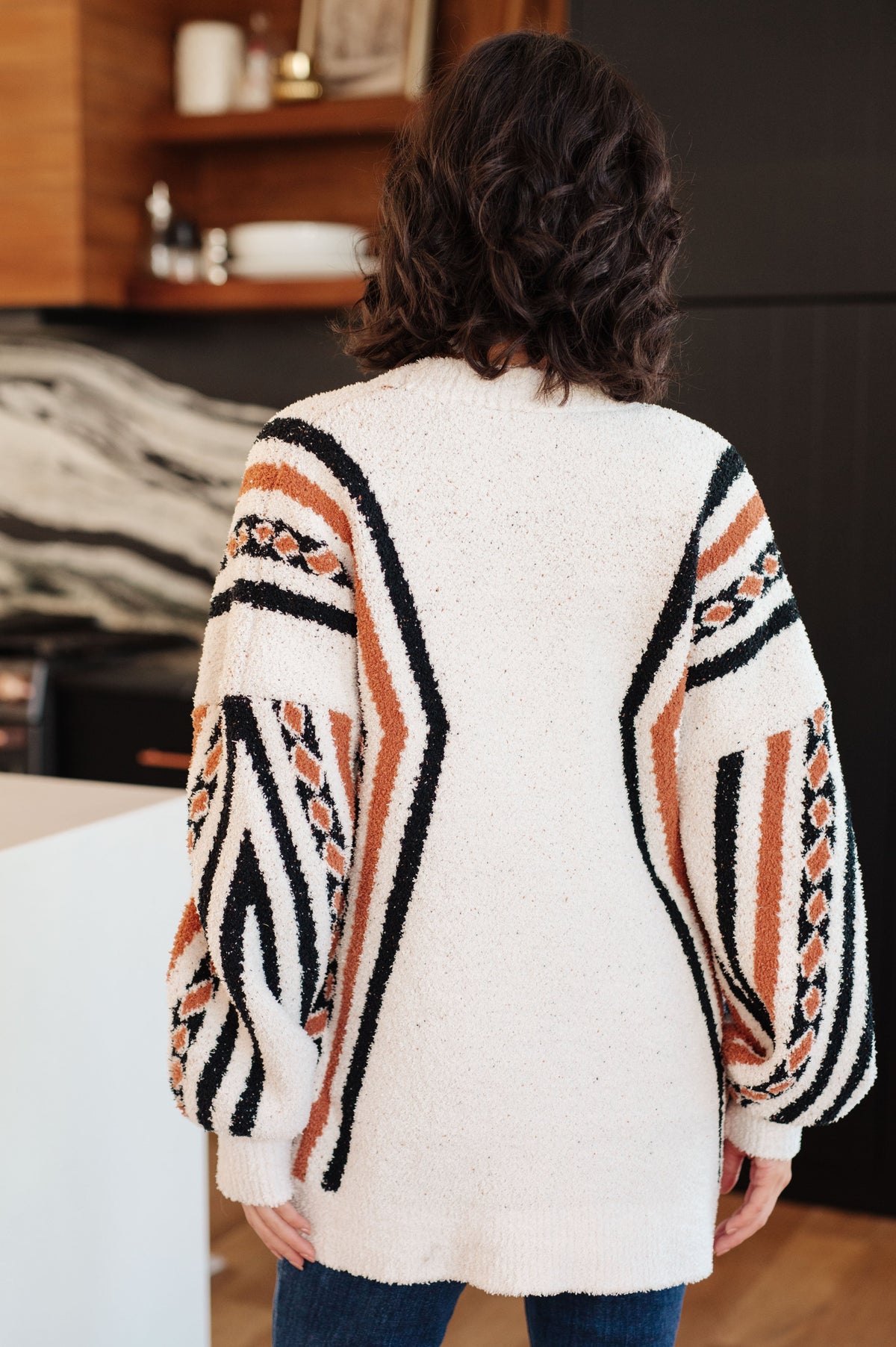 Back view of a woman in a cream fuzzy knit cardigan with bold black and orange patterned sleeves and a relaxed fit indoors