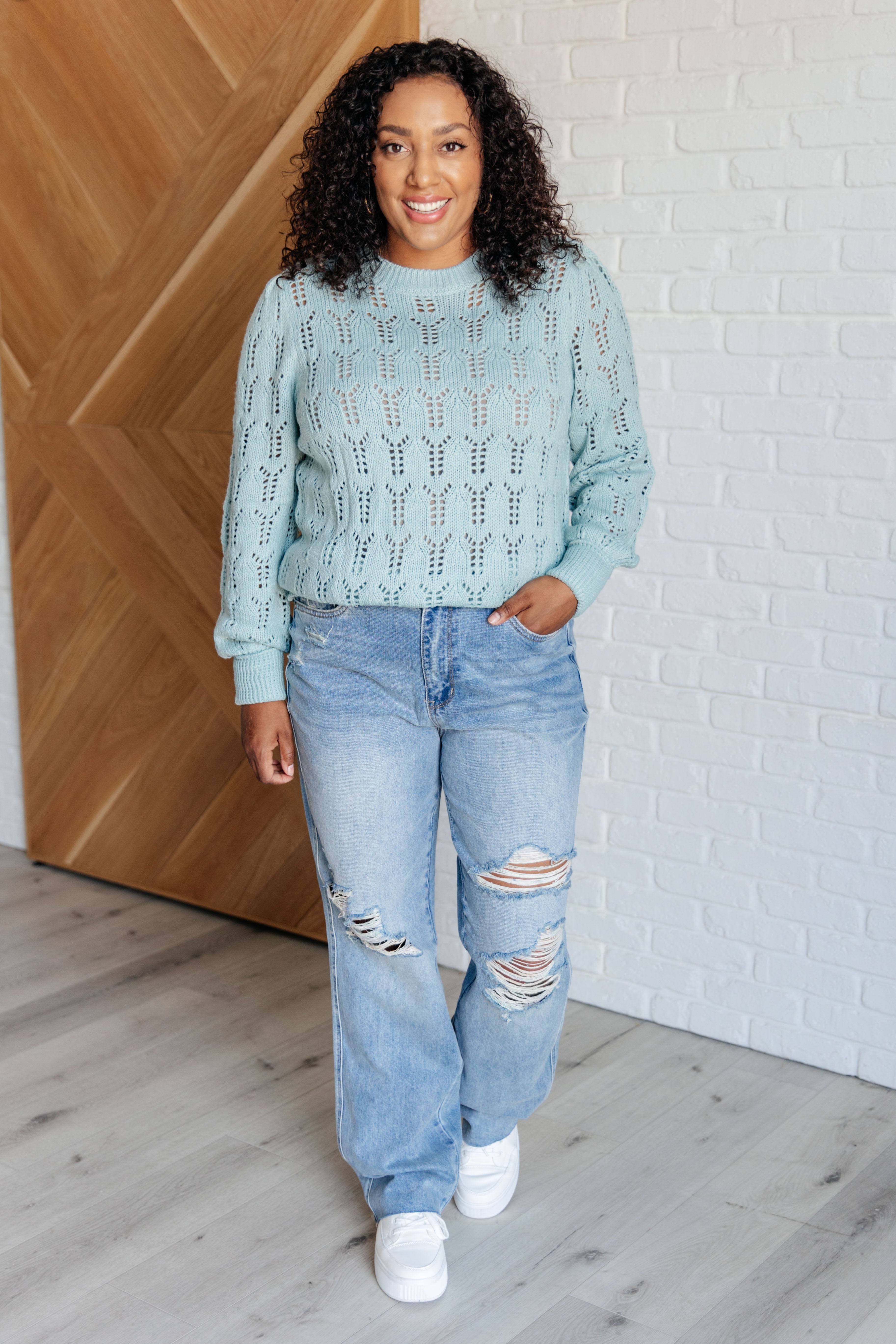 Woman smiling wearing a light blue sheer pointelle knit sweater paired with ripped light wash jeans and white sneakers