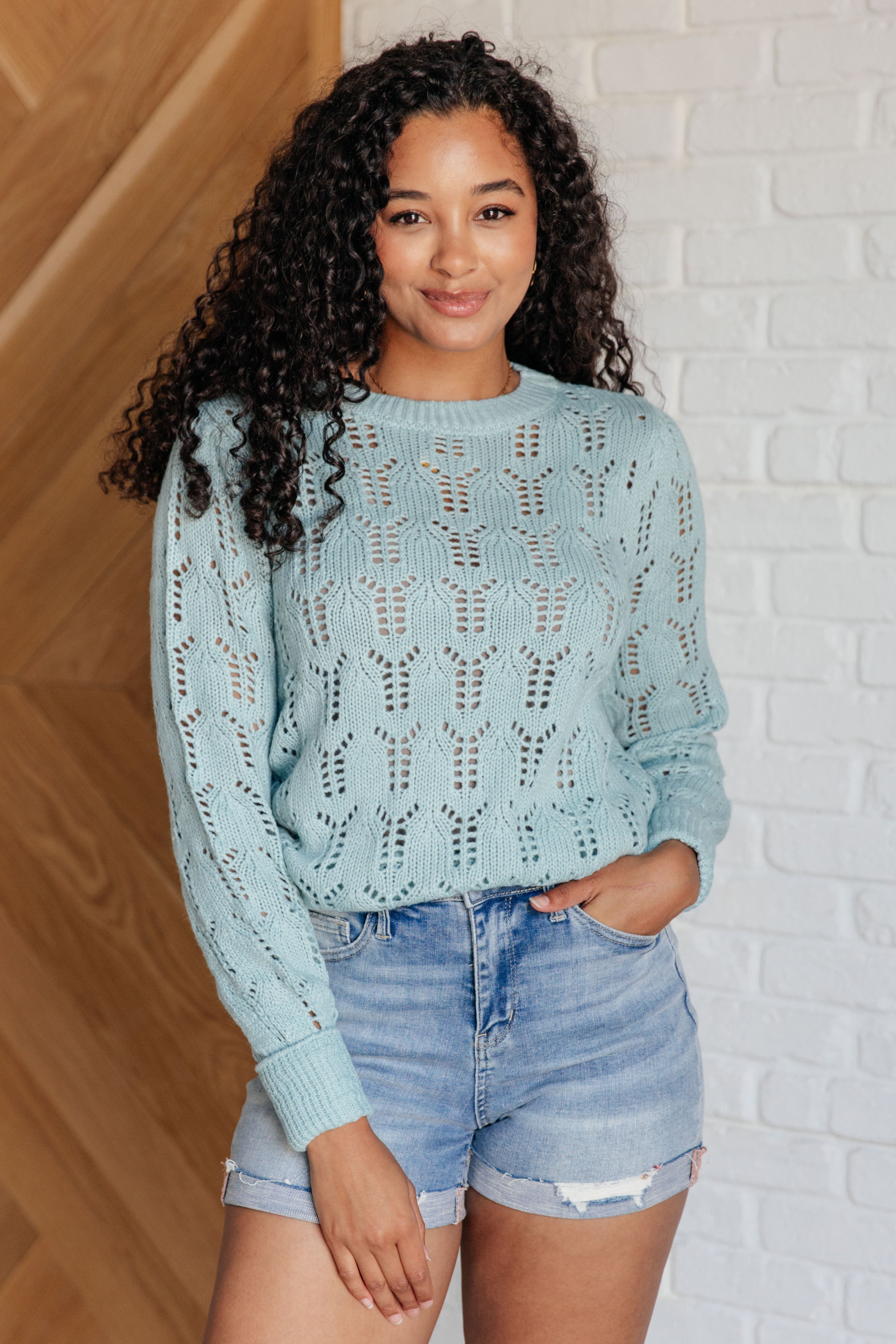 Woman wearing a light blue sheer pointelle knit sweater with long sleeves and patterned openwork paired with denim shorts