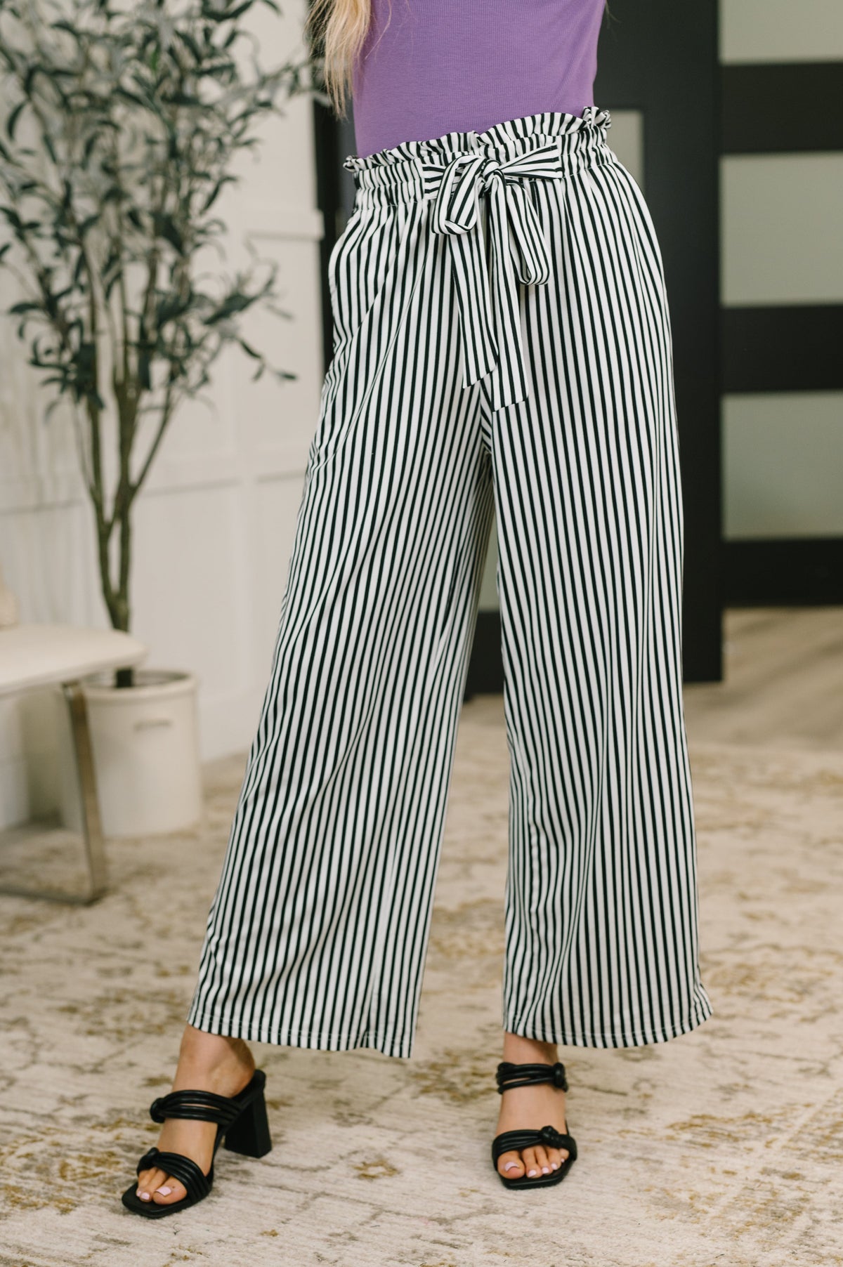 Woman wearing Black and White striped high waisted wide leg trousers with a tie waist black heeled sandals and a purple top in a home interior
