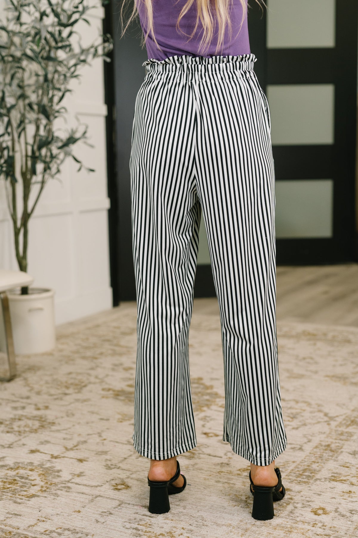Woman seen from behind in black and white striped high waisted wide leg trousers a purple top and black heeled sandals indoors