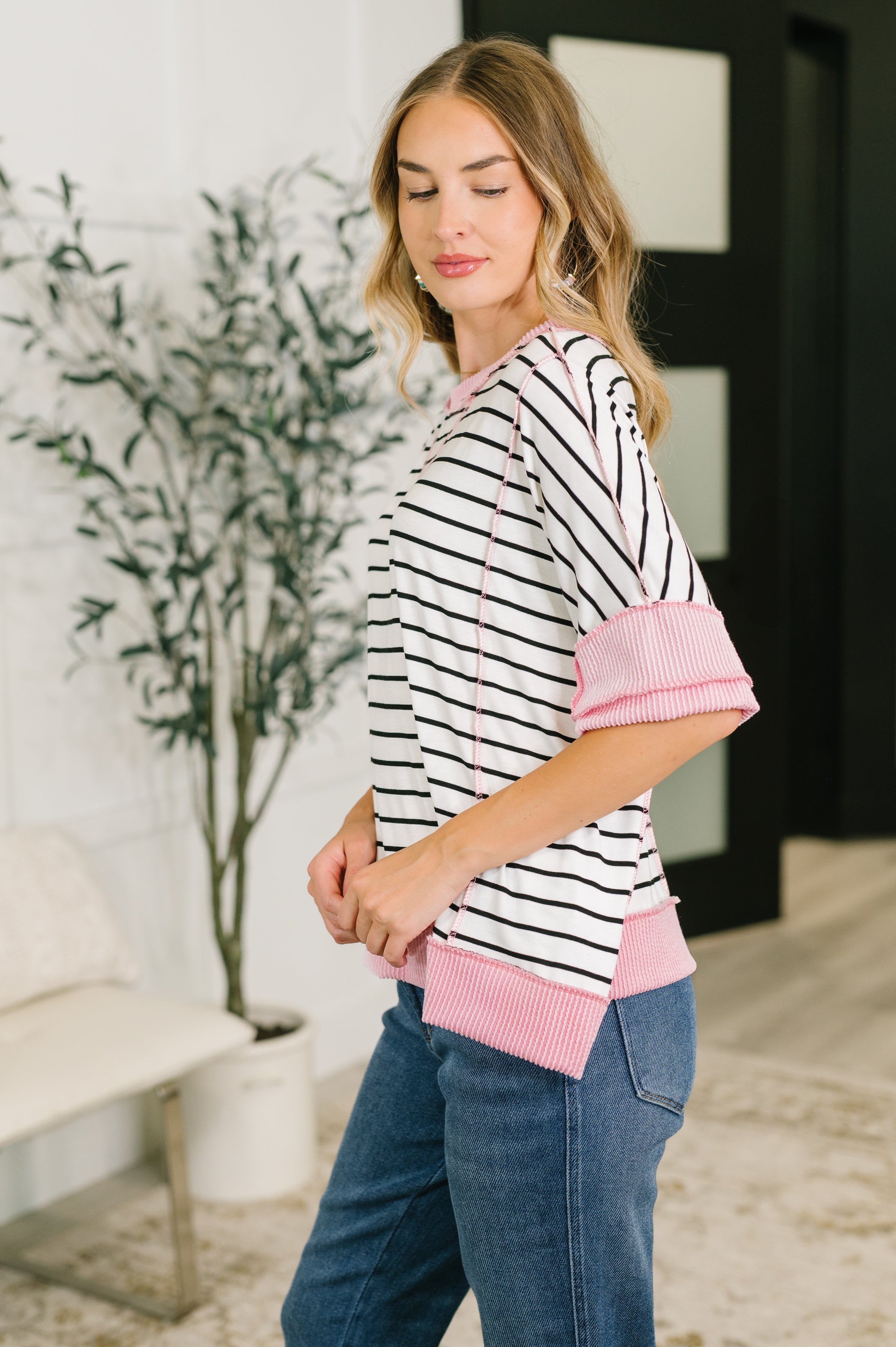 Side profile of woman in striped top with pink contrast cuffs and hem paired with blue jeans indoors