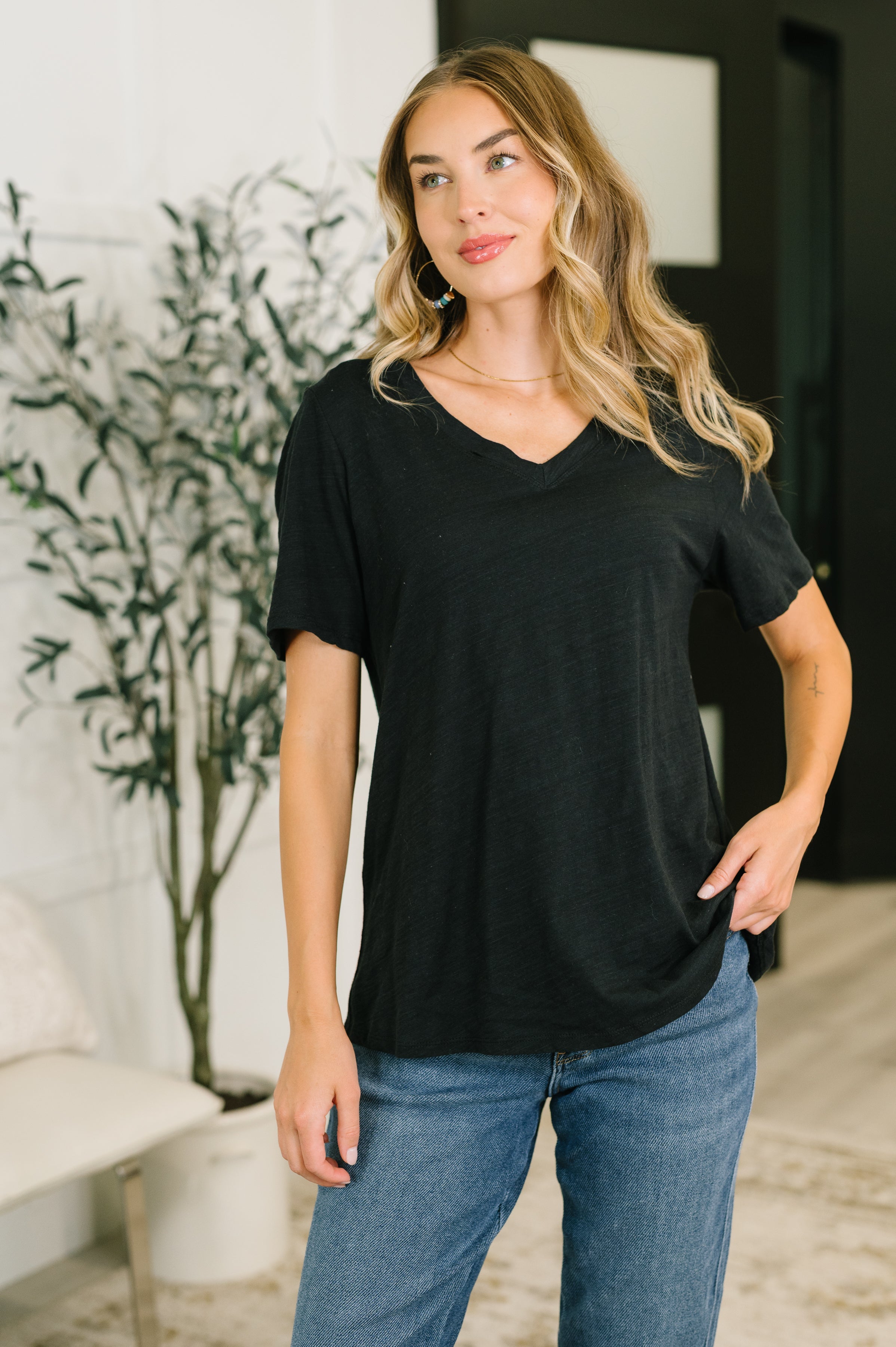 Woman wearing relaxed fit black V-neck tee with short sleeves and blue jeans indoors near plant and chair