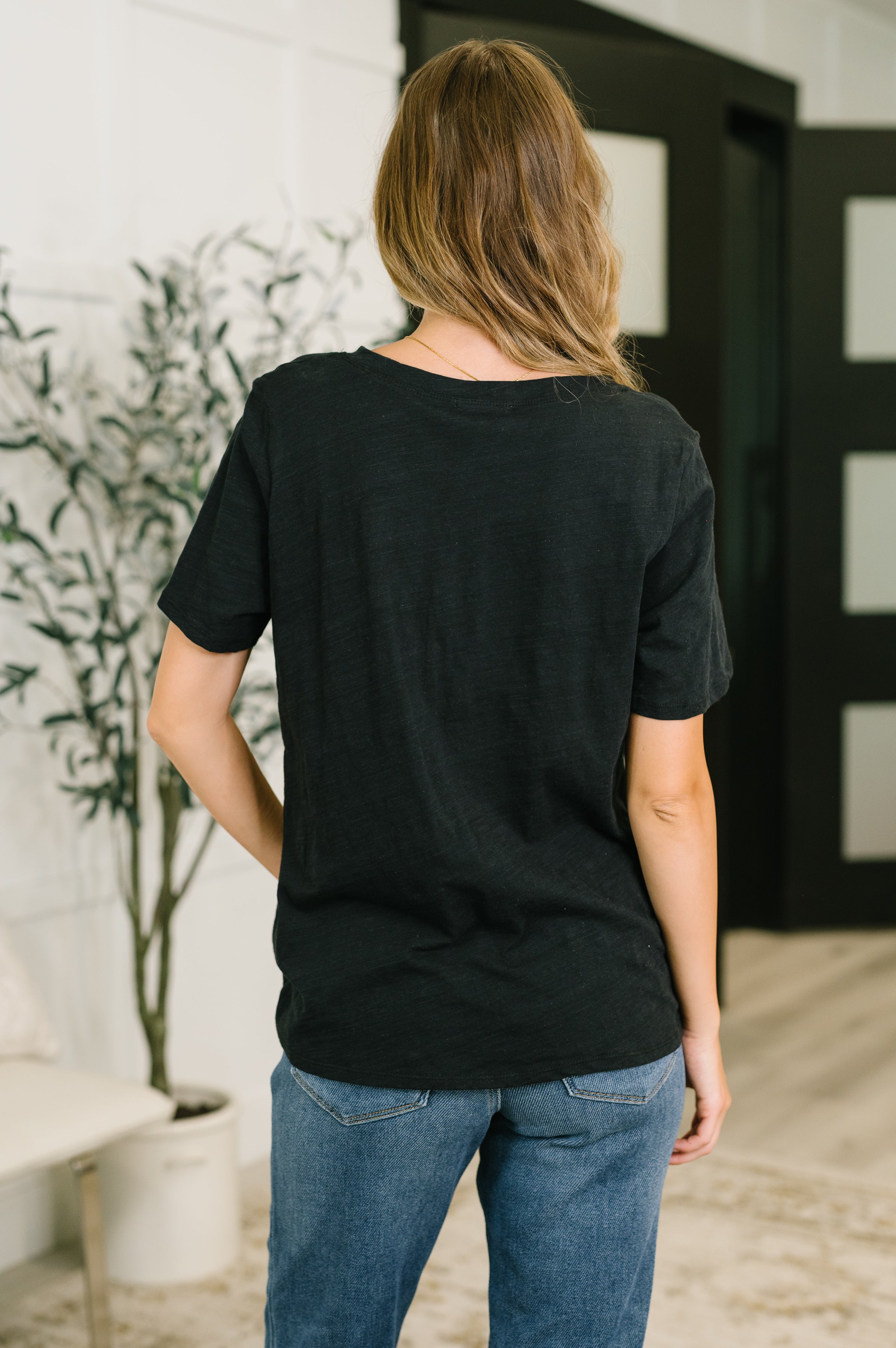 Woman wearing relaxed black short sleeve tee paired with blue jeans standing indoors with plant in background